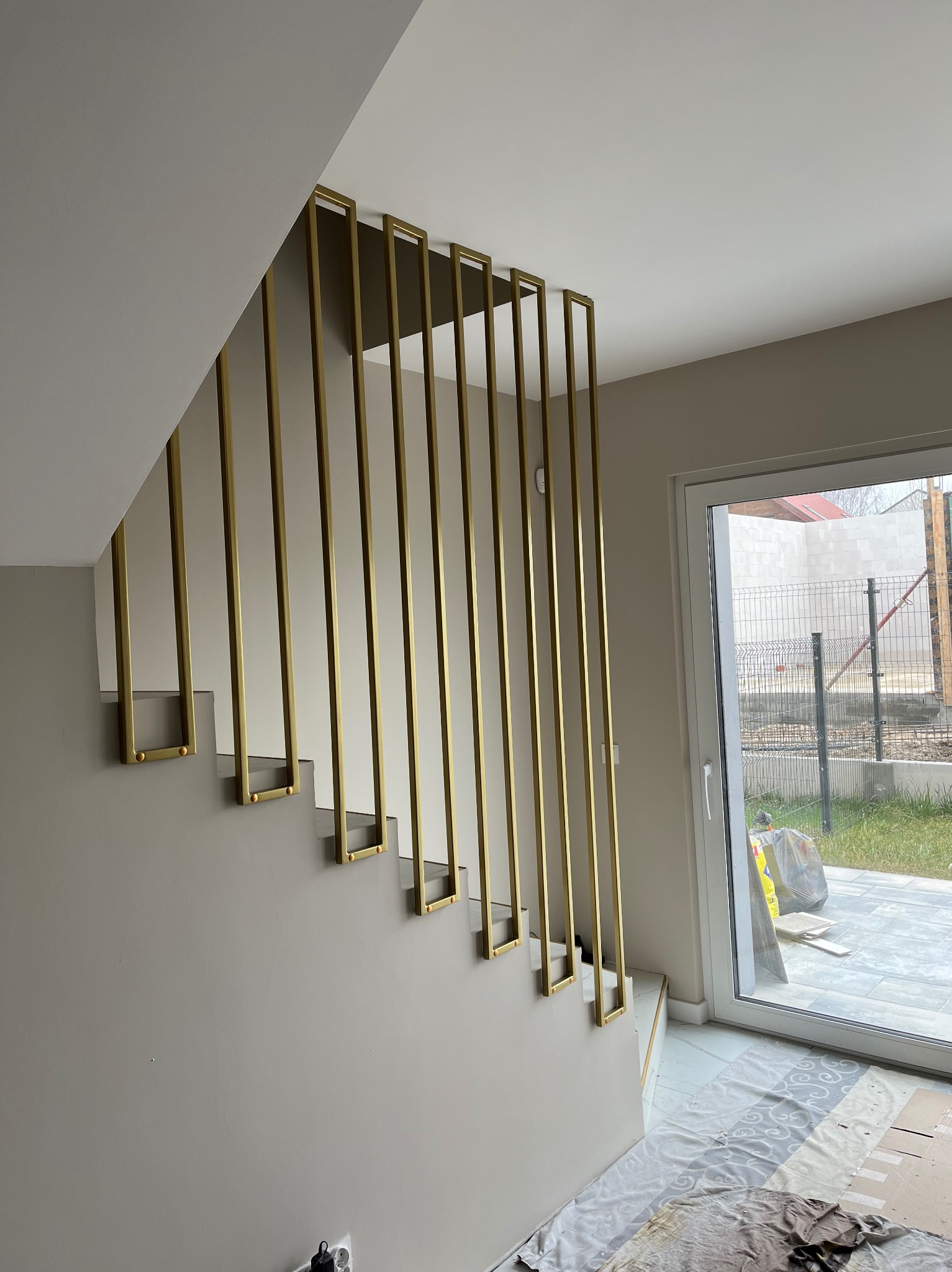Nowoczesne schody z minimalistyczną, złotą balustradą w formie pionowych, prostokątnych profili, zakończonych małymi, okrągłymi elementami mocującymi. Widok na zewnątrz przez przeszklone drzwi...