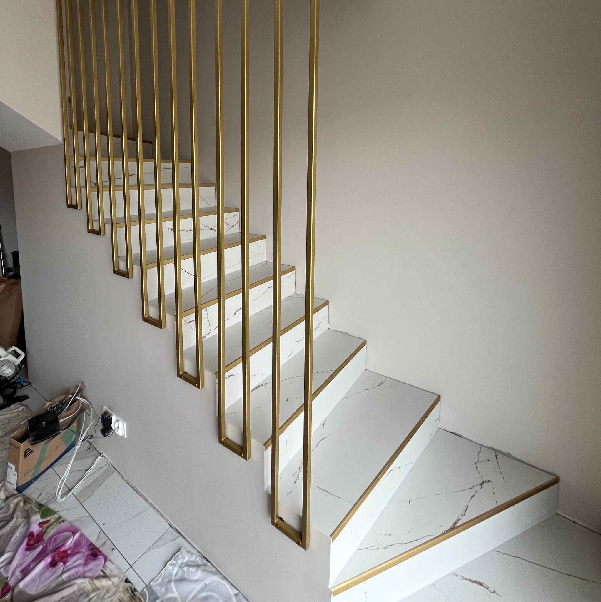 Białe schody z marmurowym wzorem i złotymi detalami, zabezpieczone pionowymi, złotymi prętami tworzącymi minimalistyczną barierkę.