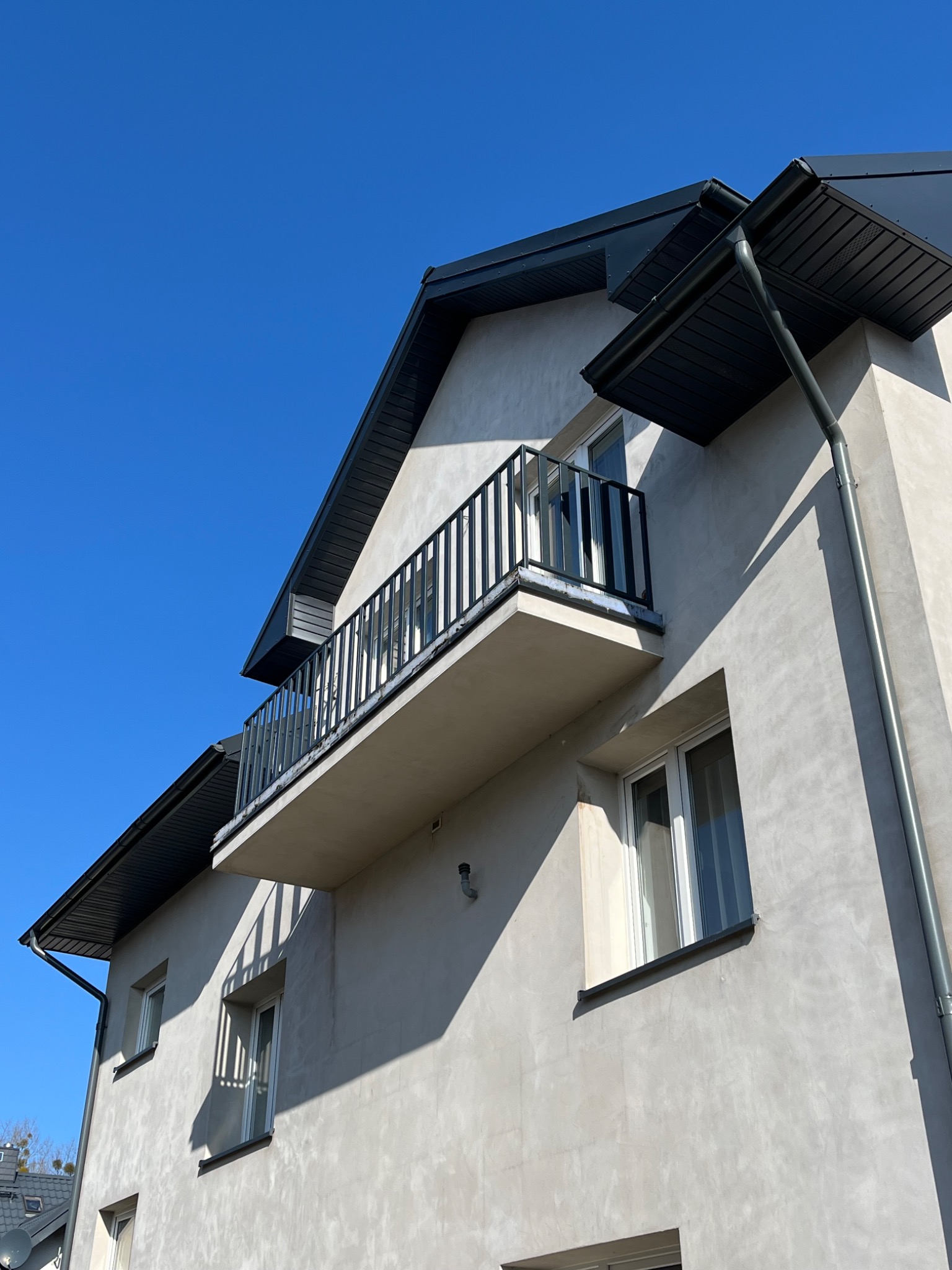 Fasada budynku z balkonem zabezpieczonym czarną, metalową balustradą wertykalną, widziana z dołu pod ostrym kątem, na tle błękitnego nieba.