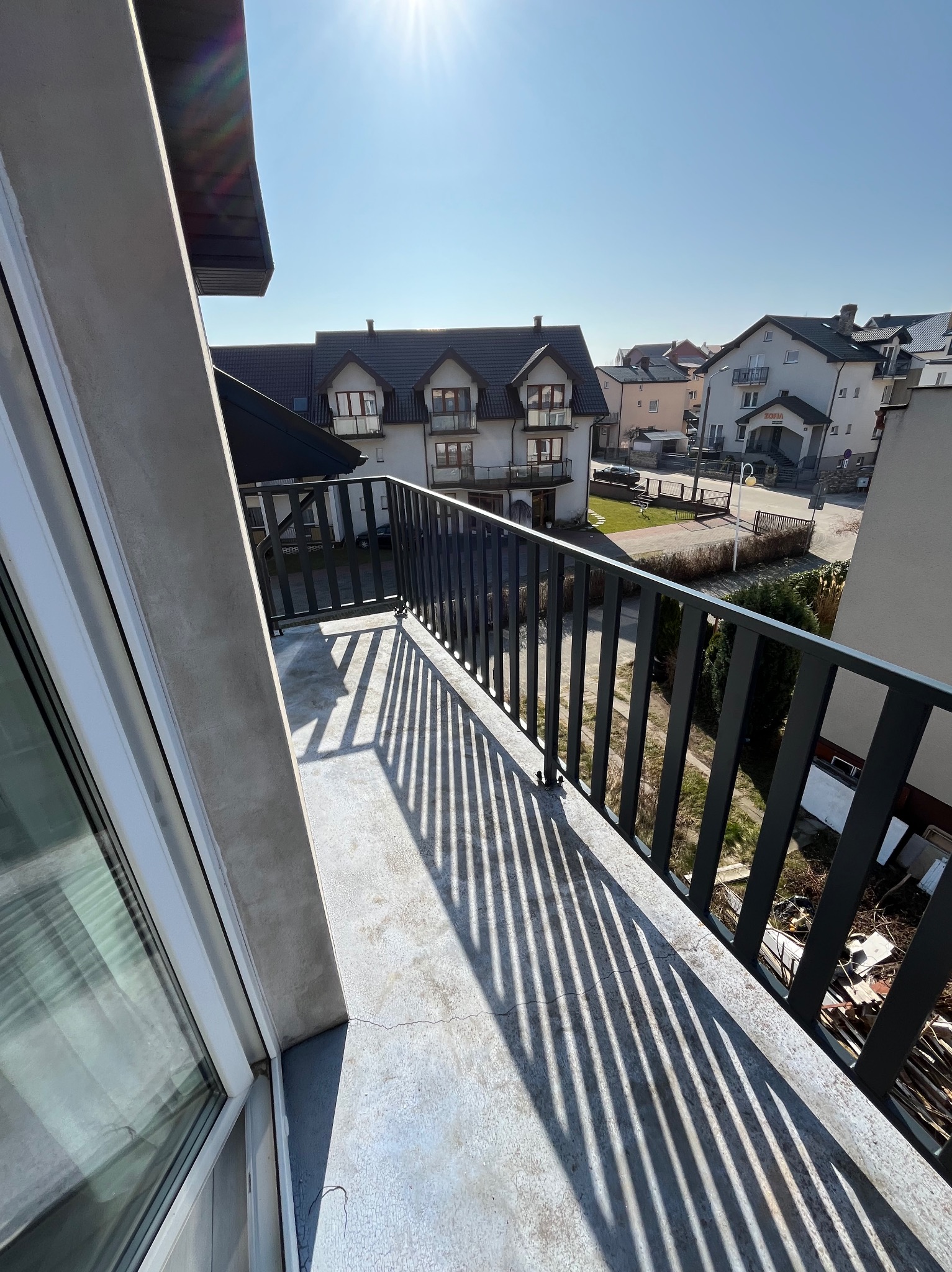 Ciemna, metalowa balustrada balkonowa rzuca wyraźne cienie na betonową podłogę balkonu w słoneczny dzień. W tle budynki mieszkalne.