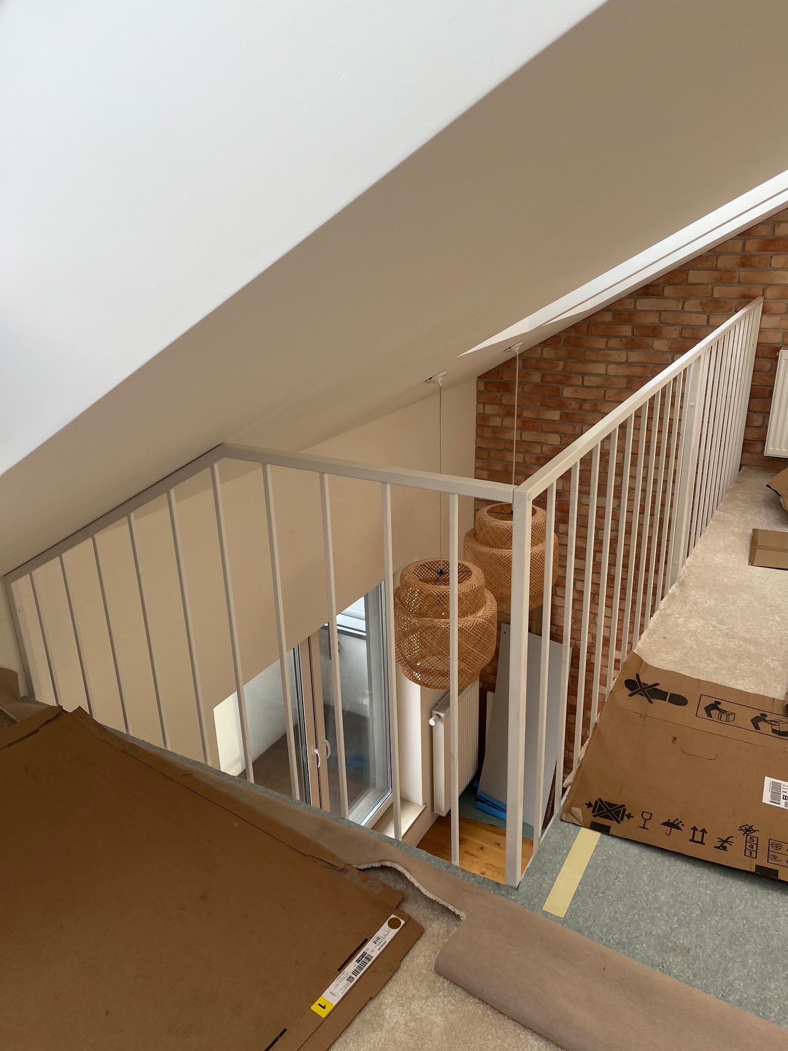 Biała, metalowa balustrada schodowa z pionowymi szczeblami w nowym domu, widok z góry na klatkę schodową, w tle ściana z czerwonej cegły i wiszące lampy z rattanu, na podłodze kartony i elementy...