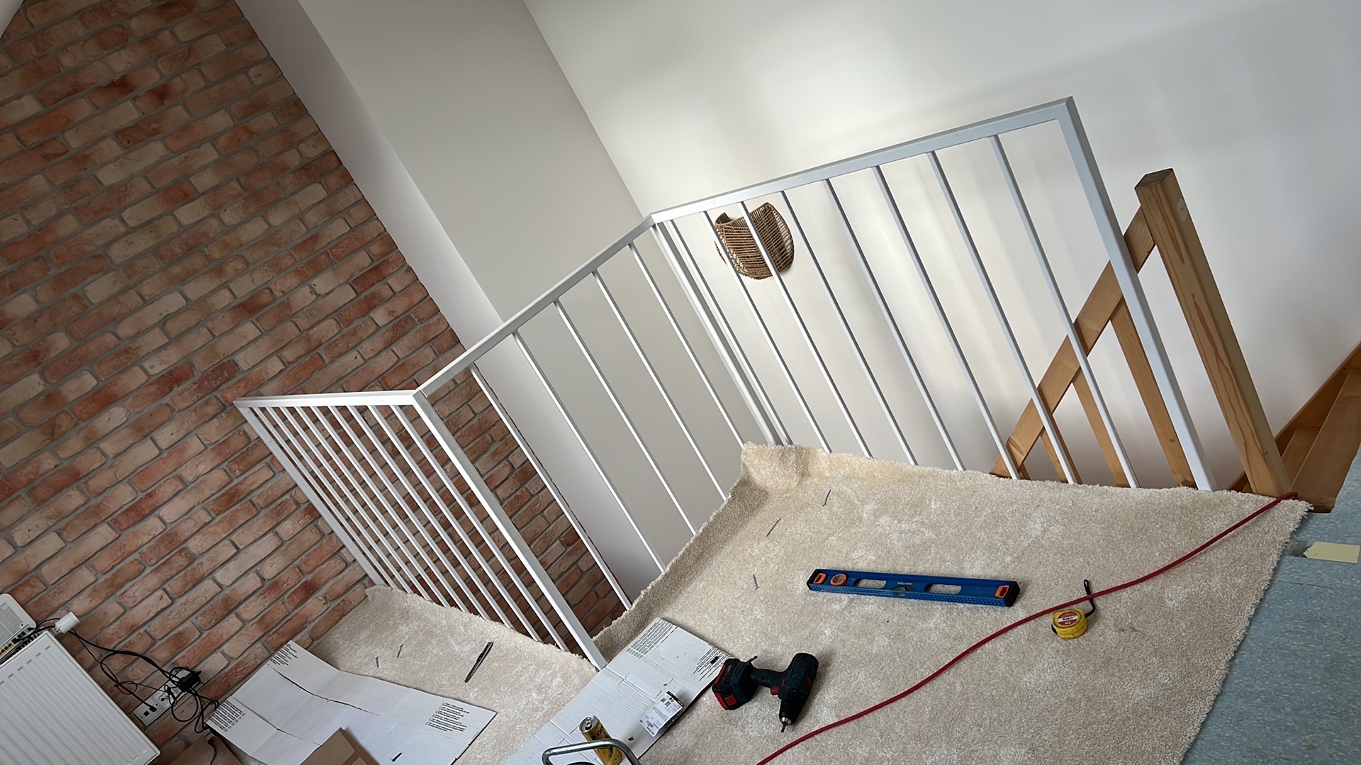 Montaż białej balustrady schodowej z pionowymi szczeblami, widoczne narzędzia: poziomica, wkrętarka, miarka, kartony z rozrysowanymi elementami, tło stanowi ściana z cegły i fragment schodów...