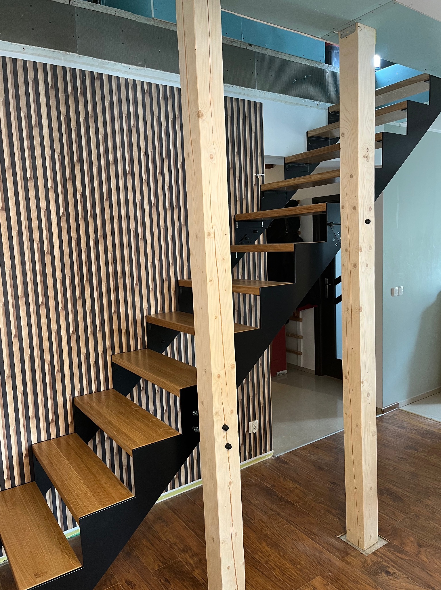 Nowoczesne schody z drewnianymi stopniami i czarną metalową konstrukcją, wsparte na drewnianych słupach, ustawione na tle ściany z dekoracyjnymi panelami w pionowe pasy.