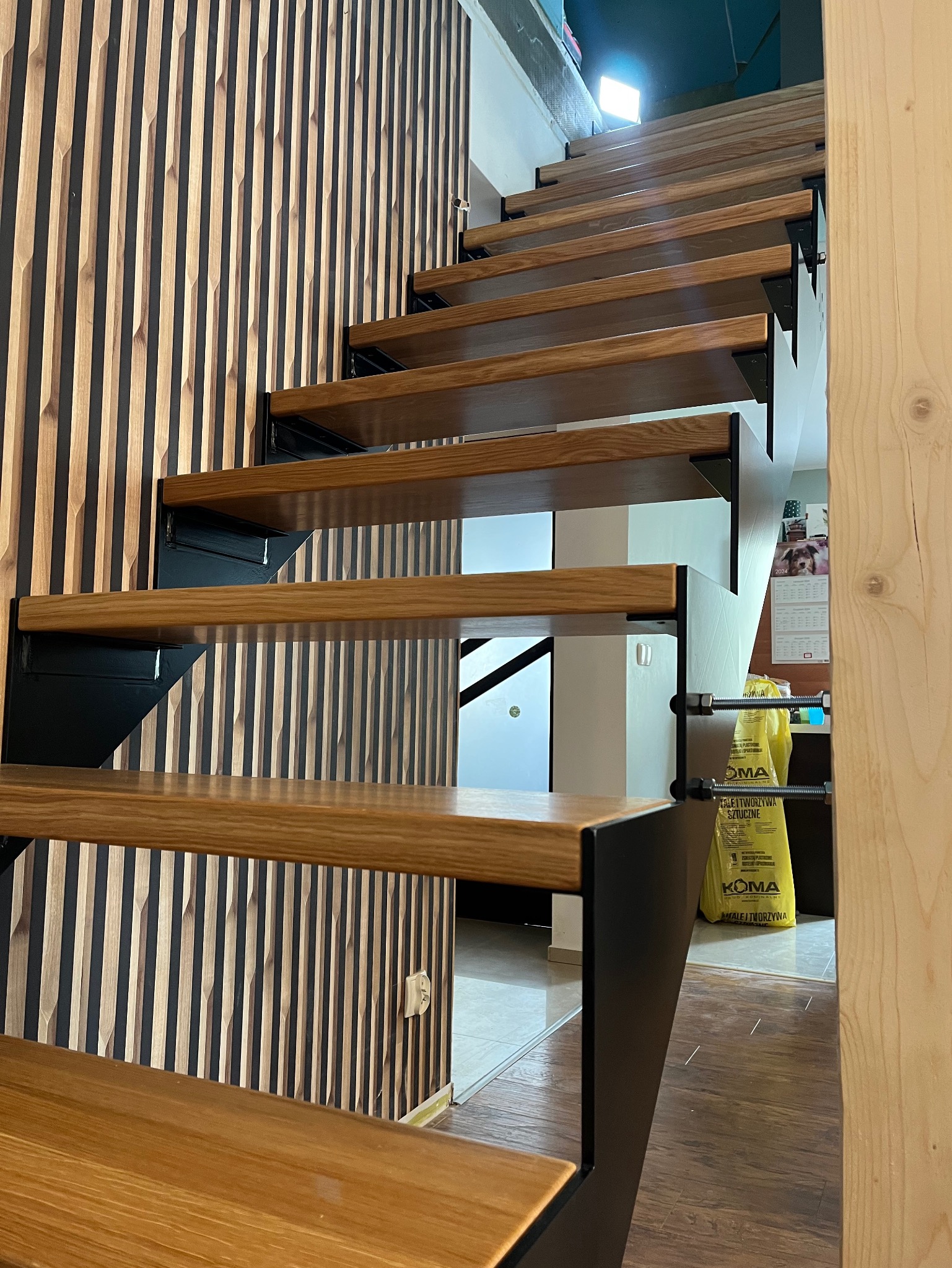 Drewniane schody metalowe z czarnymi wspornikami, widok z dołu na tle ściany z drewnianymi panelami, w tle żółte worki z tworzywa.