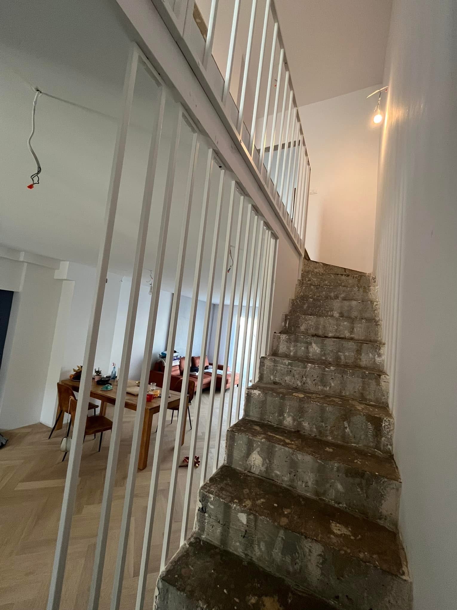 Wnętrze w trakcie remontu: surowe betonowe schody prowadzą na górę, zabezpieczone białą, minimalistyczną barierką. W tle widoczny salon z drewnianym stołem i pomarańczową sofą. Z sufitu zwisa kabel.