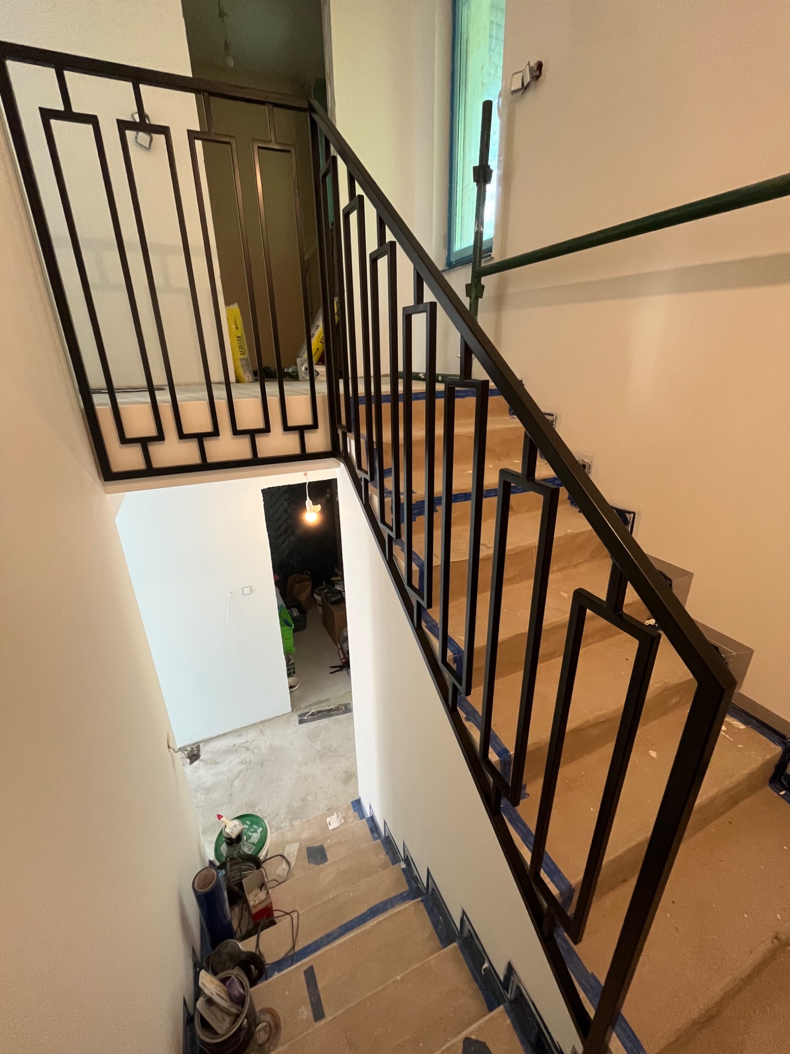 Schody w trakcie wykończenia z nowoczesną, czarną metalową konstrukcją poręczy o geometrycznym wzorze, widok z góry na narzędzia i materiały budowlane na stopniach.