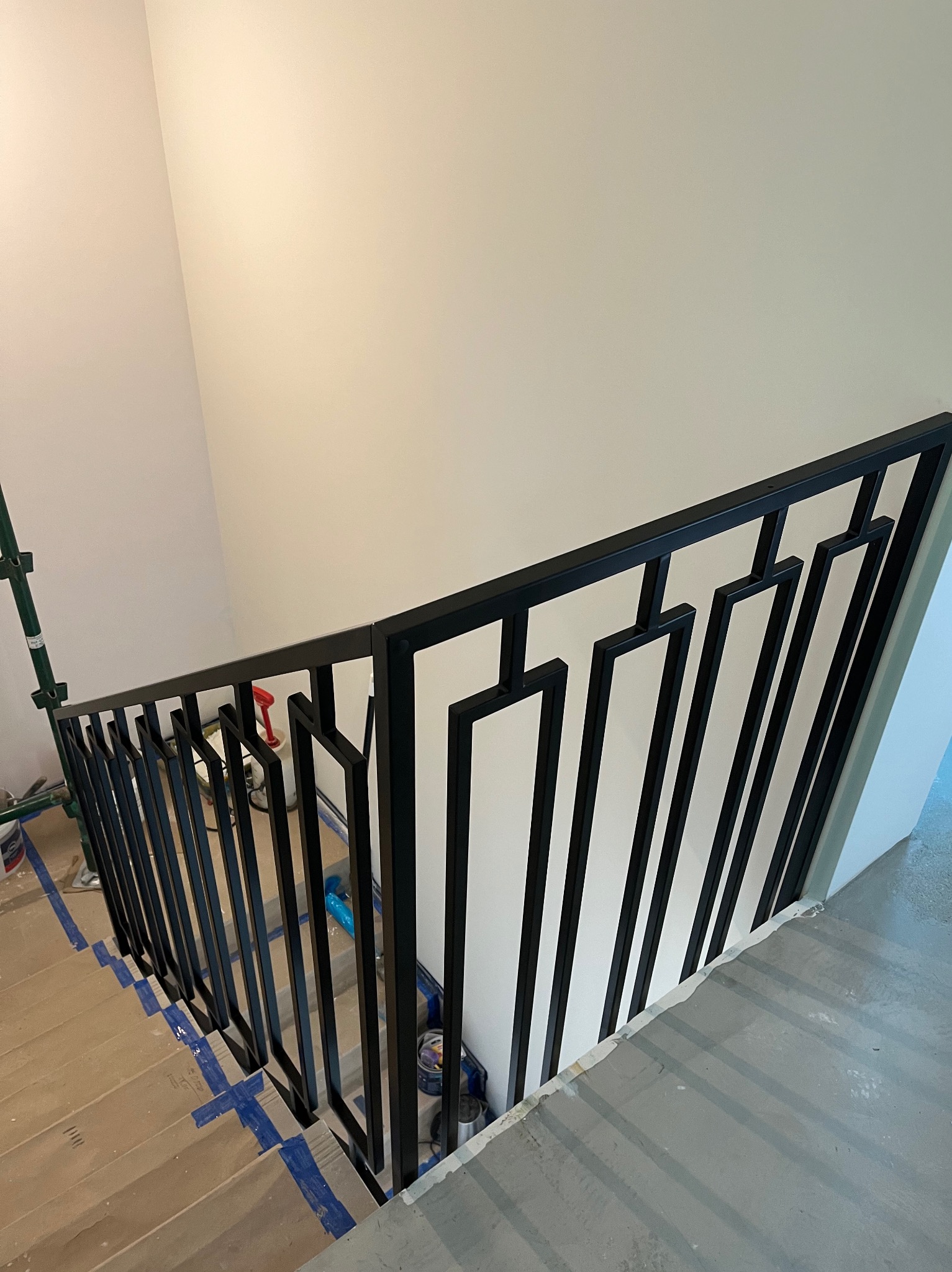 Nowoczesna, czarna barierka schodowa z geometrycznym wzorem, widoczna podczas montażu; zabezpieczone schody i ściany, w tle rusztowanie malarskie.