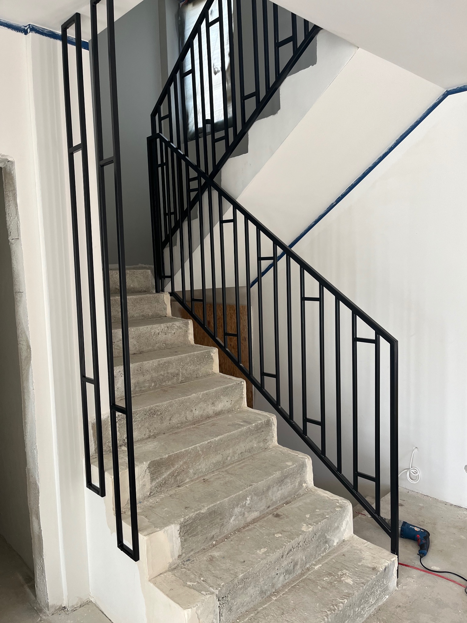 Surowe betonowe schody z nowoczesną, czarną, metalową balustradą o minimalistycznym designie, widoczne ślady prac wykończeniowych na ścianach i podłodze, w tle okno z zasłoniętą szybą i narzędzie...