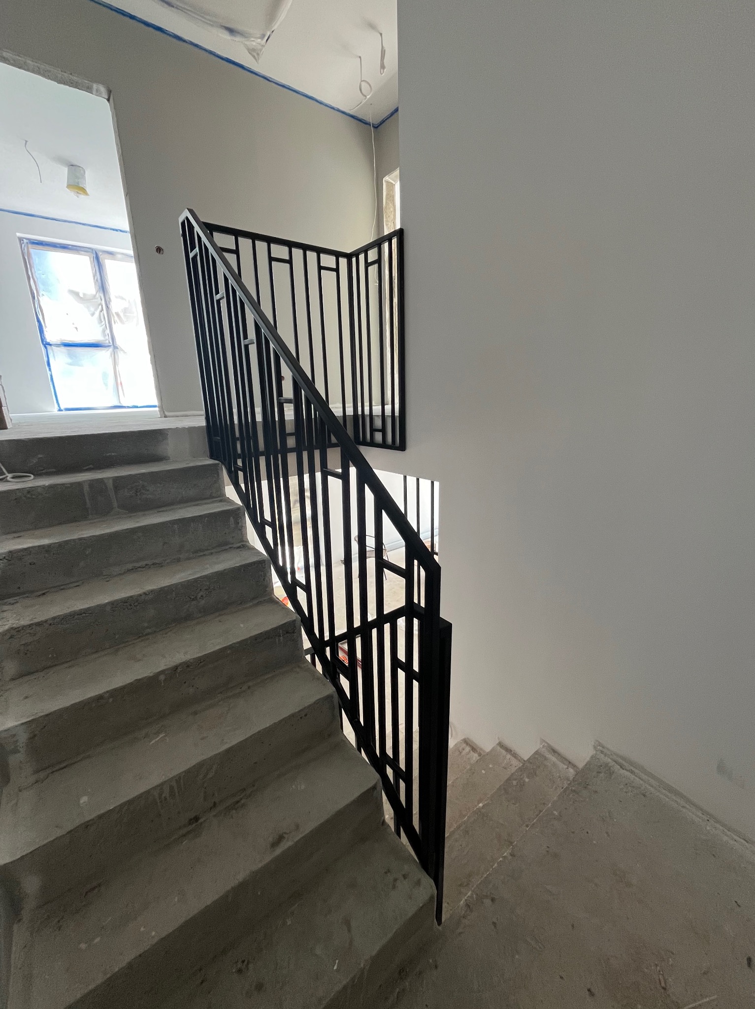 Surowe betonowe schody z czarną, metalową balustradą o minimalistycznym designie, w trakcie prac wykończeniowych, z widocznymi zabezpieczeniami okien i ścian.