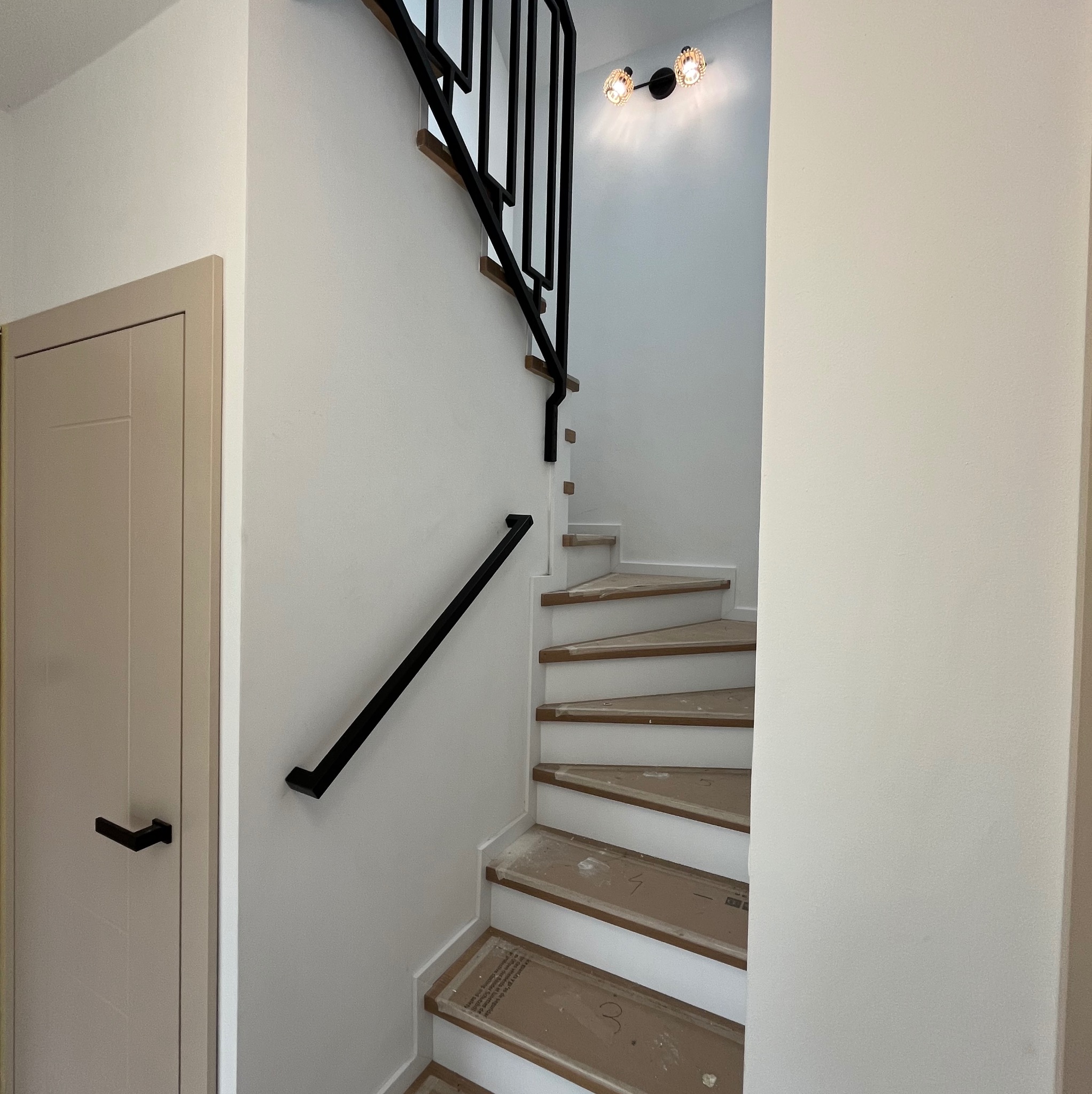 Wnętrze domu w trakcie wykańczania: schody z drewnianymi stopniami zabezpieczonymi kartonem, czarna, minimalistyczna poręcz i balustrada z pionowych elementów, oświetlenie punktowe na ścianie.