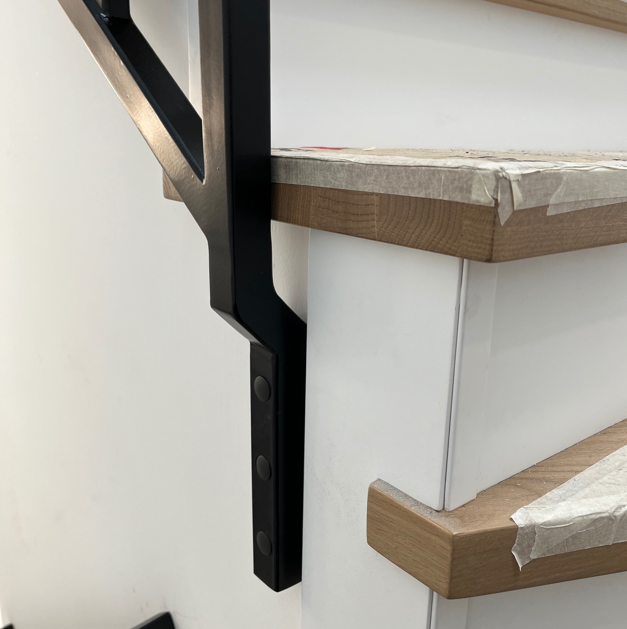 Detal czarnej, metalowej konstrukcji wspierającej drewniane stopnie schodów z białymi podstopnicami, zabezpieczone taśmą malarską podczas prac wykończeniowych.