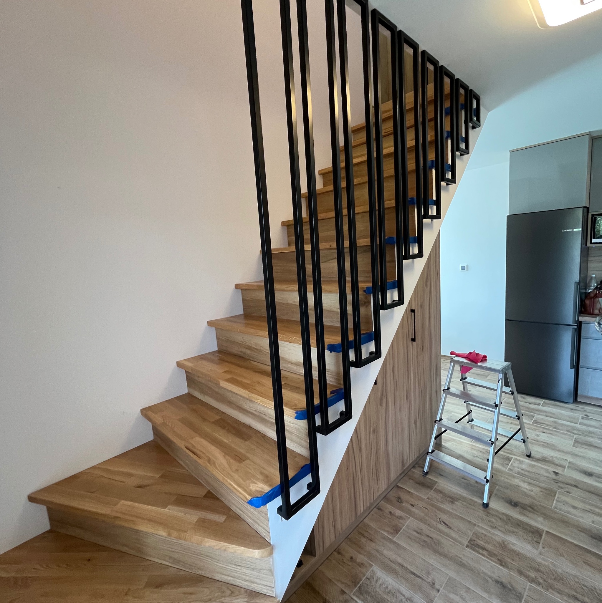 Drewniane schody z czarnymi, pionowymi słupkami balustrady, zabezpieczone niebieską taśmą malarską, widok z dołu na tle jasnej ściany i zabudowy pod schodami, z widoczną drabinką i lodówką w tle.