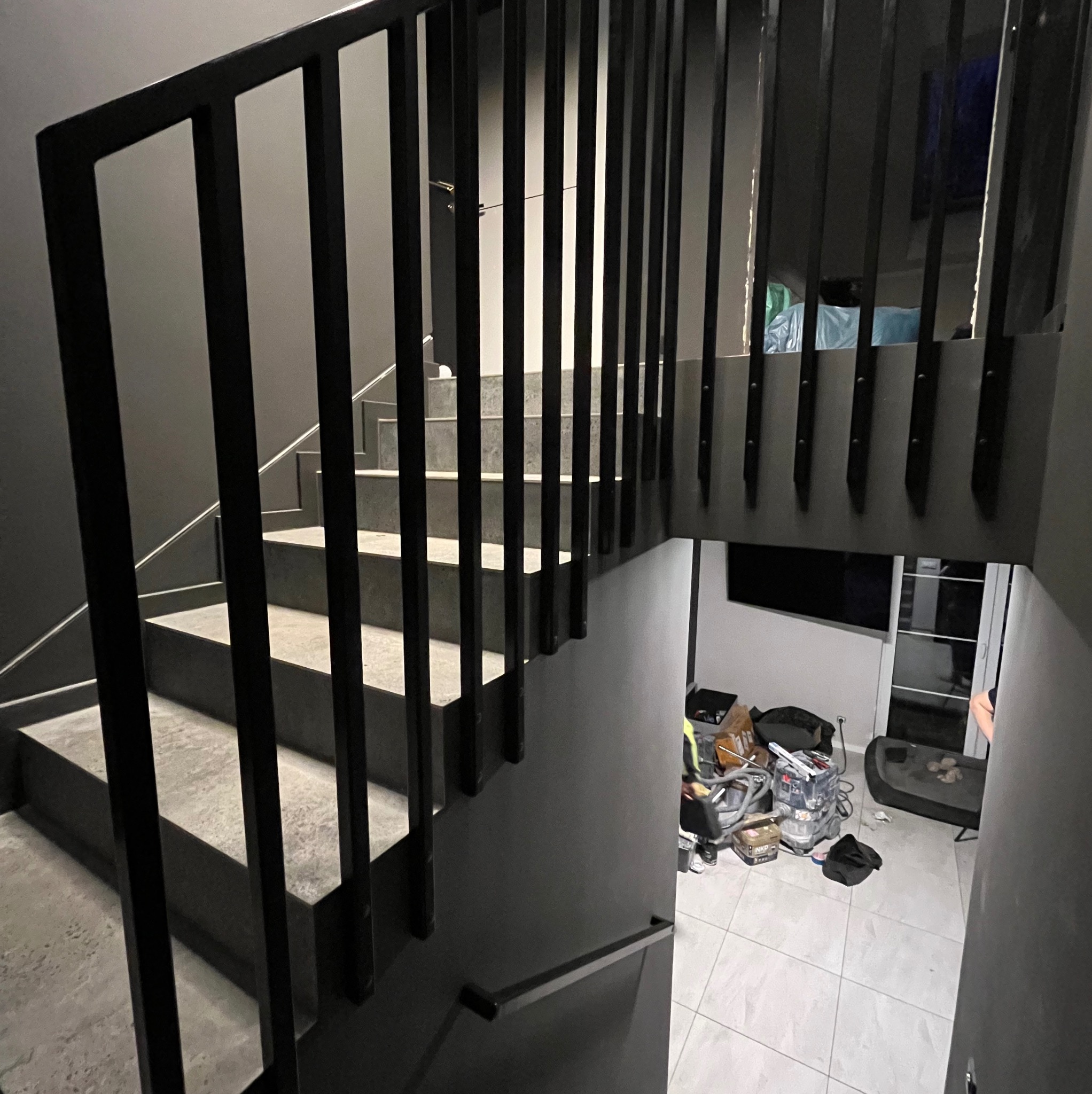 Nowoczesne schody z czarną, metalową balustradą o minimalistycznym designie, widoczne z perspektywy dolnych stopni, z fragmentem pomieszczenia z wyposażeniem widocznym w tle.