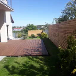 Nowoczesny taras z drewna egzotycznego przylegający do domu, otoczony trawnikiem i drewnianym ogrodzeniem lamelowym, w tle zabudowa miejska.