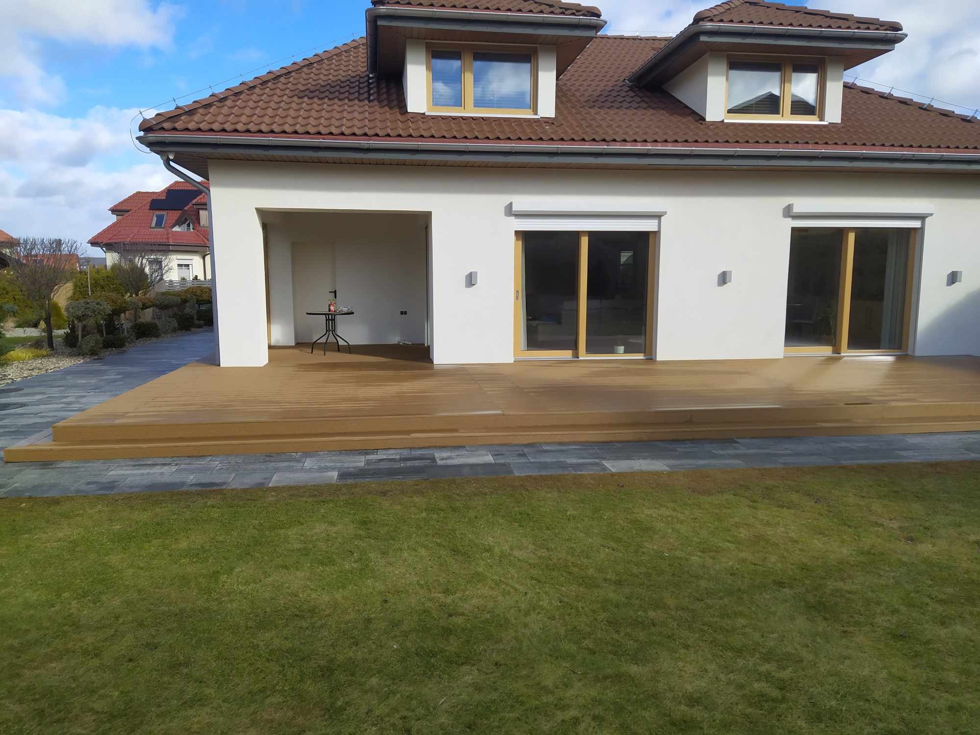 Nowoczesny dom z tarasem kompozytowym w kolorze jasnego drewna, widok z trawnika, na tarasie stolik kawowy.