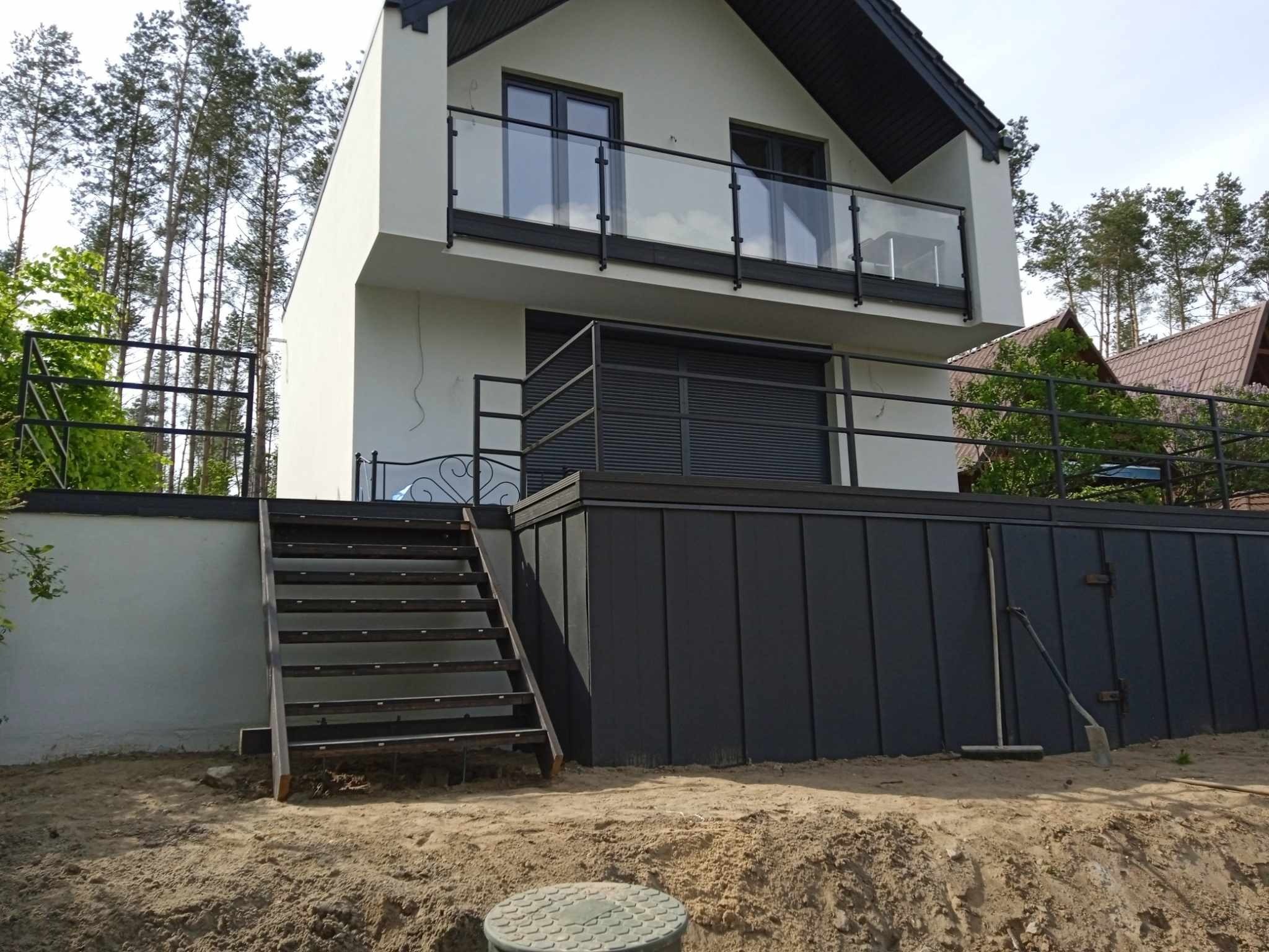 Nowoczesny taras z czarną metalową balustradą i schodami, przylegający do białego domu z balkonem, widok z poziomu gruntu na placu budowy z widoczną studzienką kanalizacyjną i narzędziami.