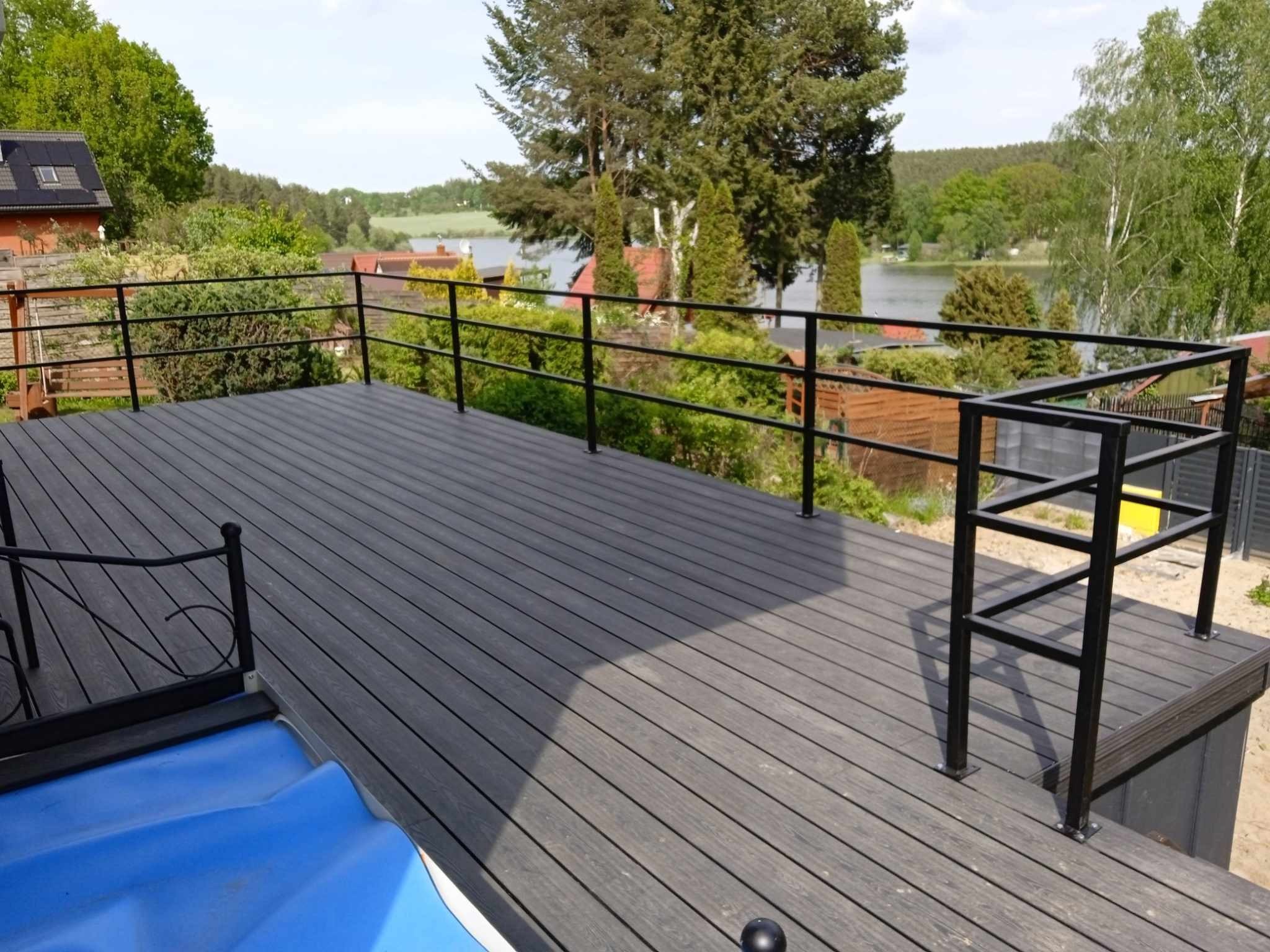 Przestronny taras kompozytowy z czarną, metalową balustradą na tle jeziora i zieleni, widoczne elementy konstrukcyjne i wykończeniowe.