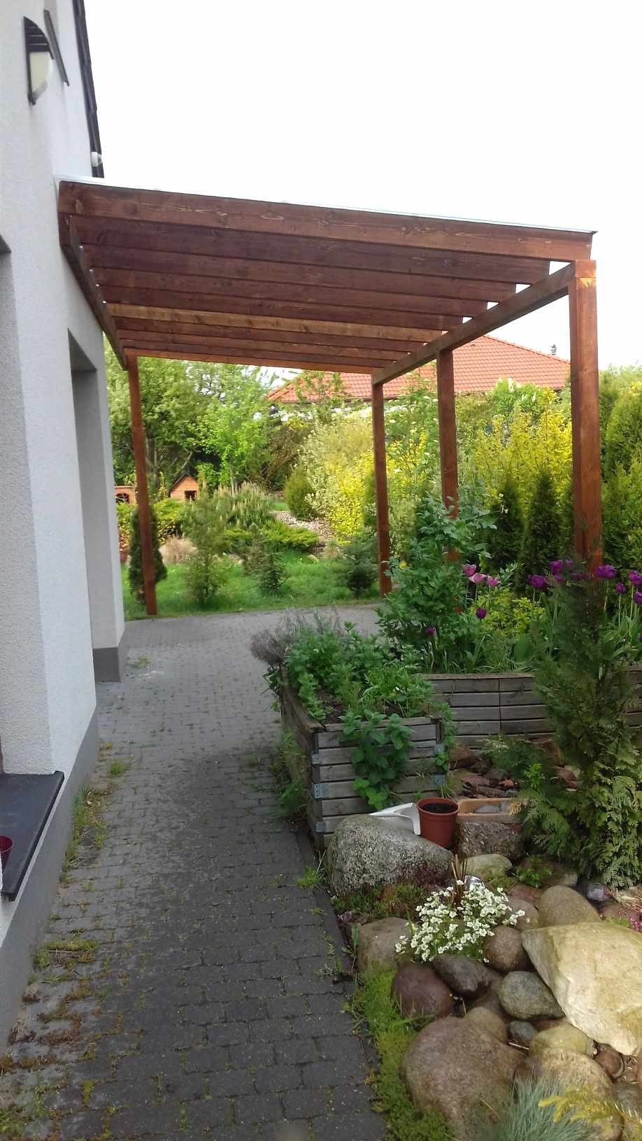 Drewniana konstrukcja zadaszenia tarasu przy domu, widok na ogród z roślinami w drewnianych skrzyniach i kamieniami dekoracyjnymi wzdłuż ścieżki z kostki brukowej.