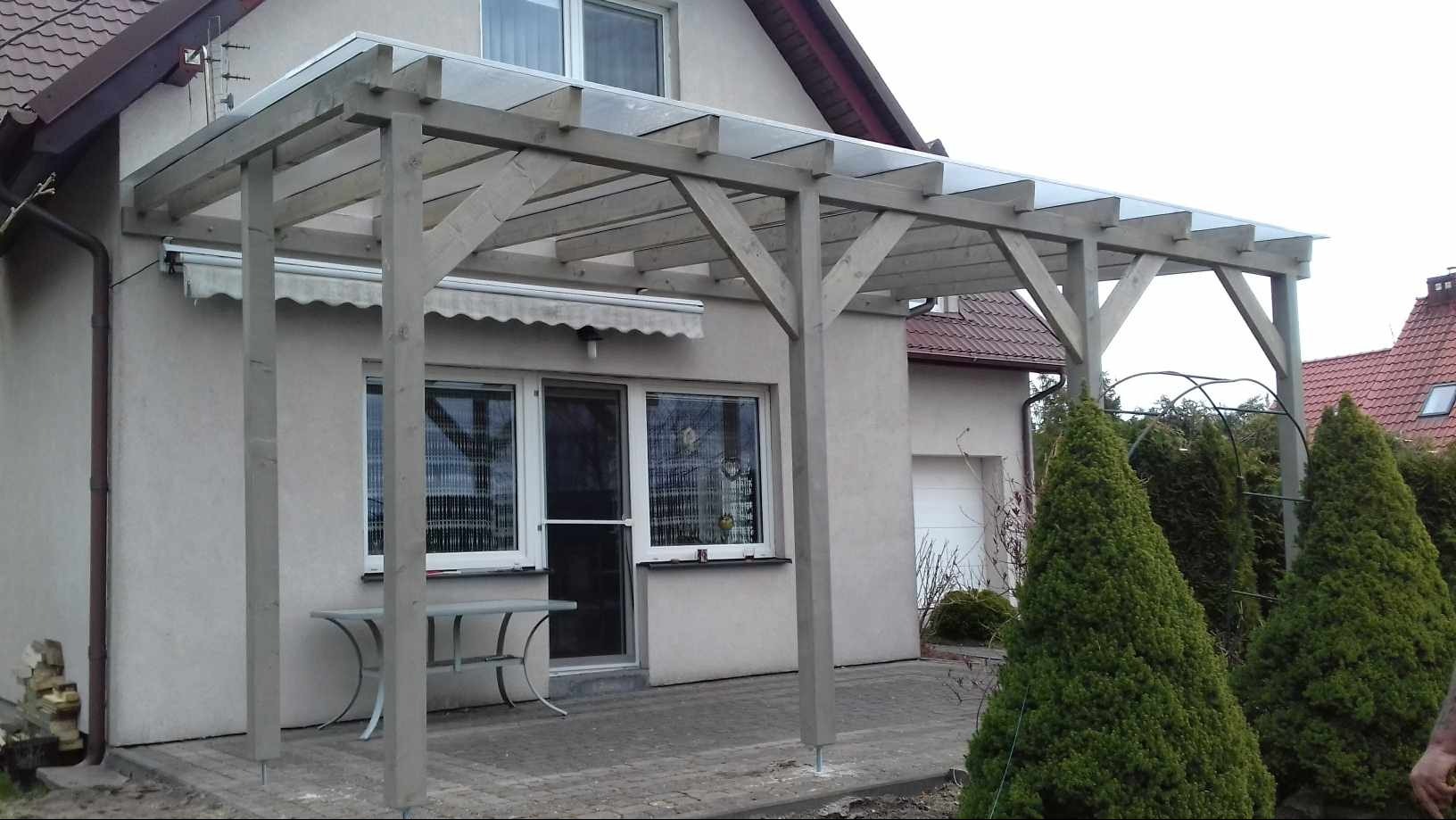 Drewniana konstrukcja z przezroczystym zadaszeniem przylegająca do budynku, z widocznym stołem i krzesłami oraz ozdobnymi iglakami w tle.