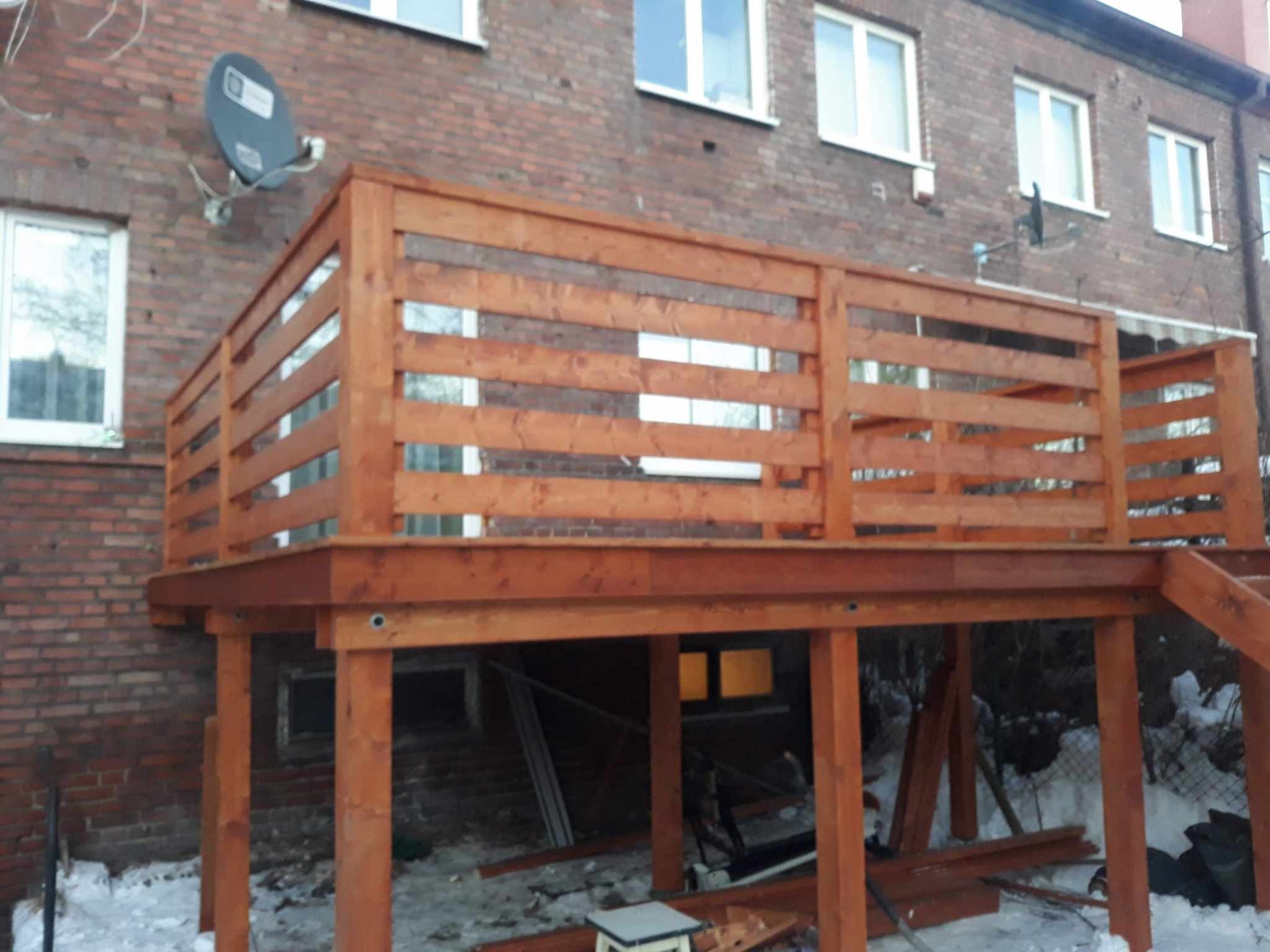 Nowy, drewniany taras przylegający do budynku z cegły, widoczny w zimowej scenerii z resztkami śniegu na ziemi.