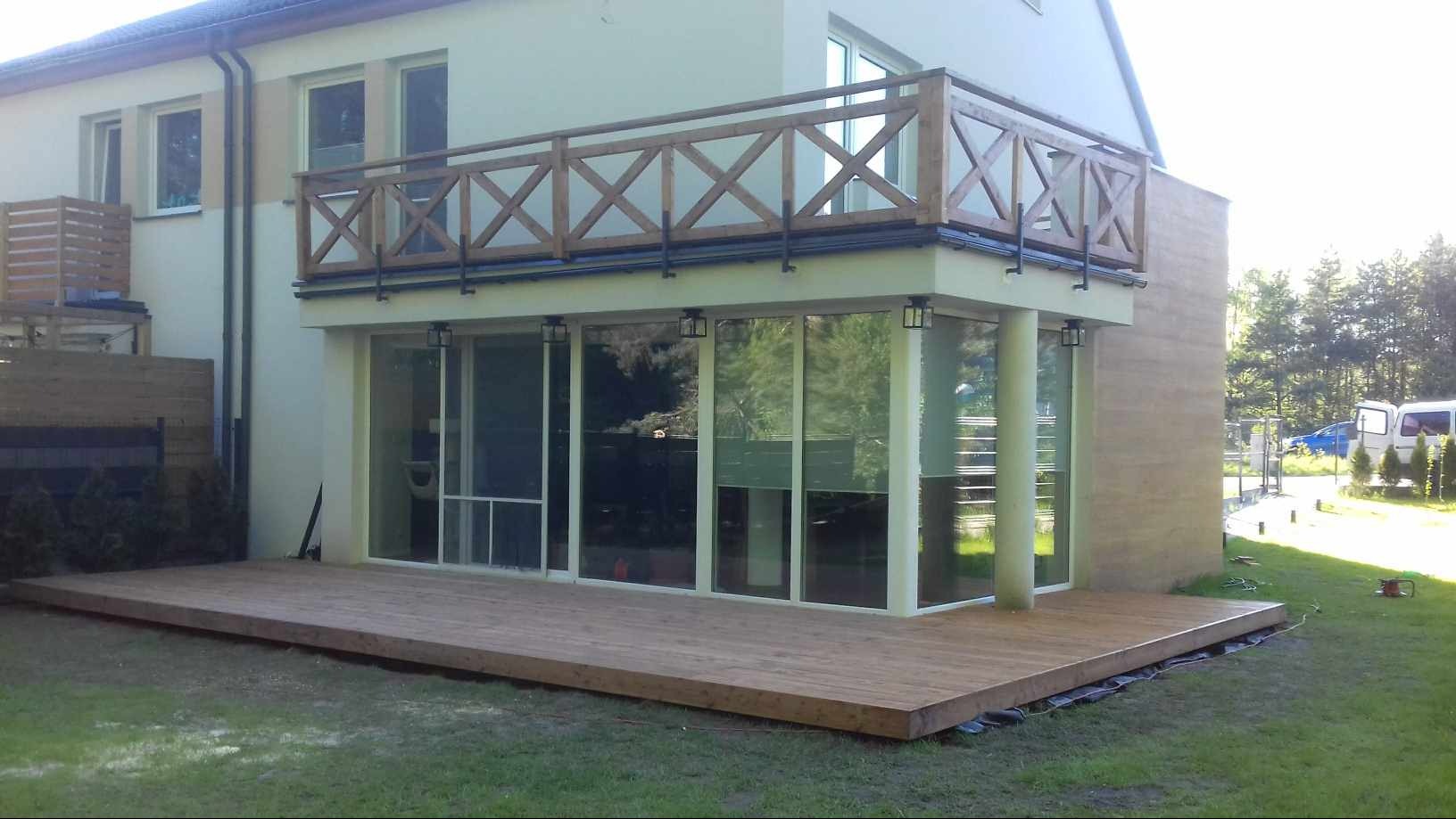 Drewniany taras przylegający do domu z dużymi oknami i balkonem z drewnianą balustradą w kształcie krzyżyków, widoczne przewody pod tarasem.