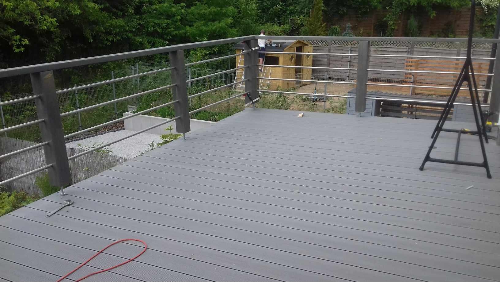 Szary taras kompozytowy z metalową balustradą w trakcie montażu, widoczny majsterkowicz na drabinie przy domku narzędziowym w tle.