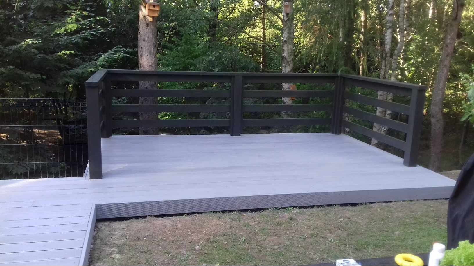 Nowoczesny taras kompozytowy w odcieniach szarości, otoczony zielenią, z ciemną balustradą i widocznymi budkami dla ptaków na drzewach w tle.