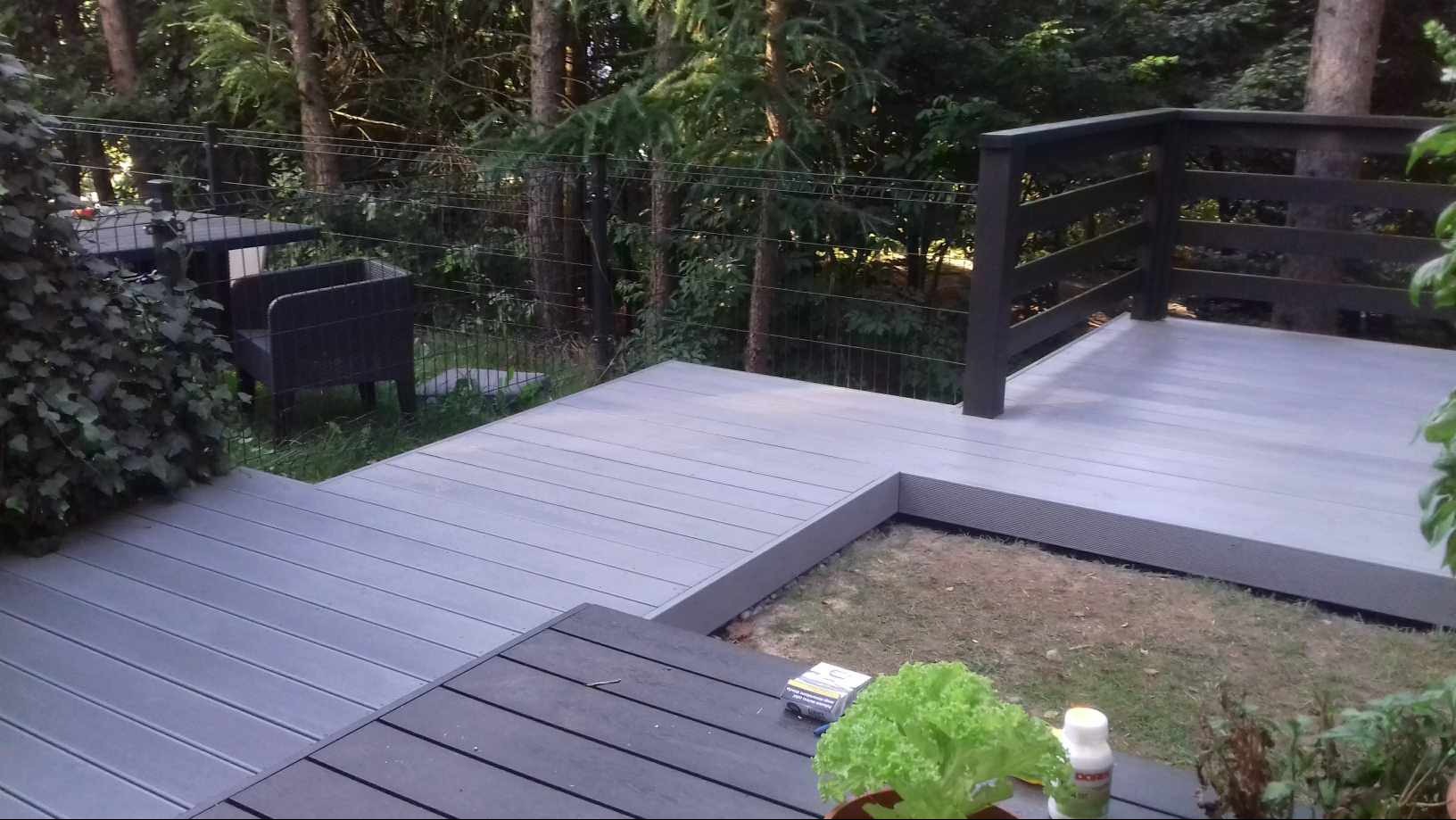 Kompozytowy taras w odcieniach szarości, z ciemną balustradą i meblami ogrodowymi w tle, otoczony zielenią drzew.