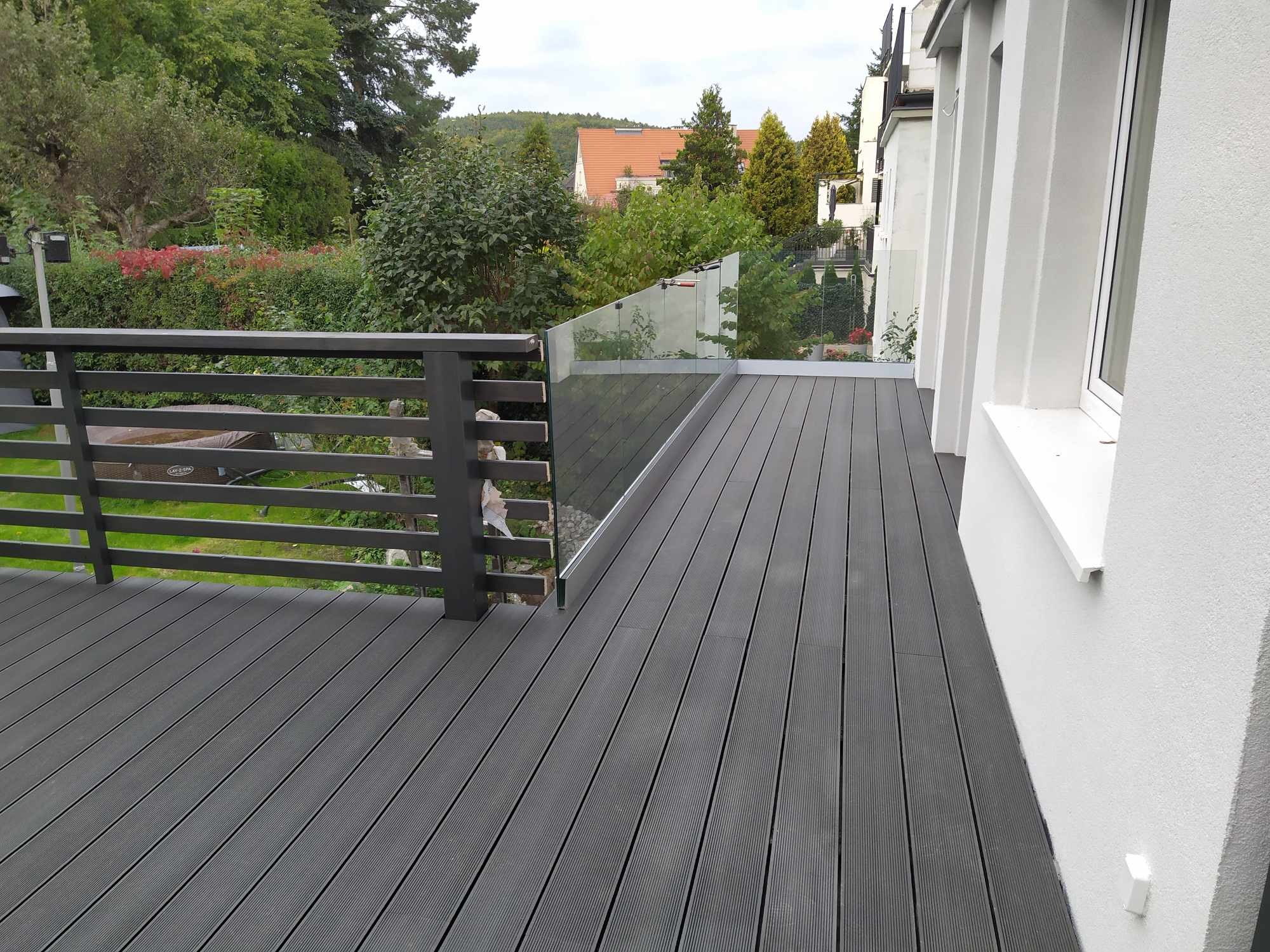 Nowoczesny taras z deski kompozytowej w odcieniach szarości, zabezpieczony szklaną balustradą i drewnianą barierką, z widokiem na zieleń ogrodu.