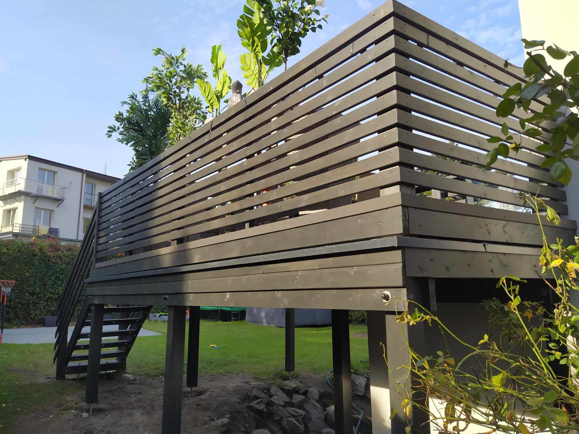 Nowoczesny taras na podwyższeniu z drewnianą balustradą z poziomych desek, schody z boku, widok z dołu na tle trawnika i domu.