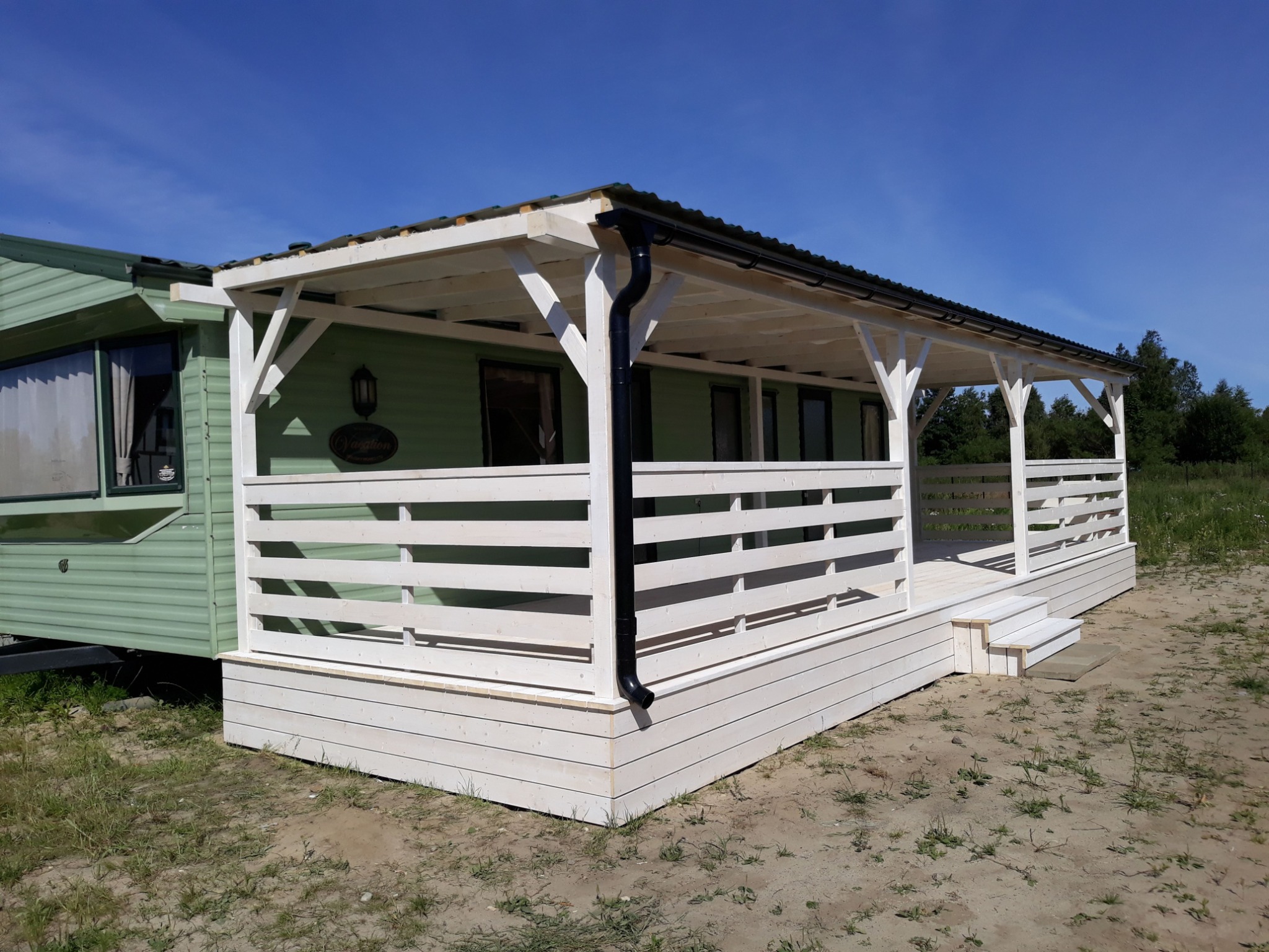 Biały taras drewniany z balustradą, przylegający do zielonego domku mobilnego z czarną rynną, na tle błękitnego nieba.