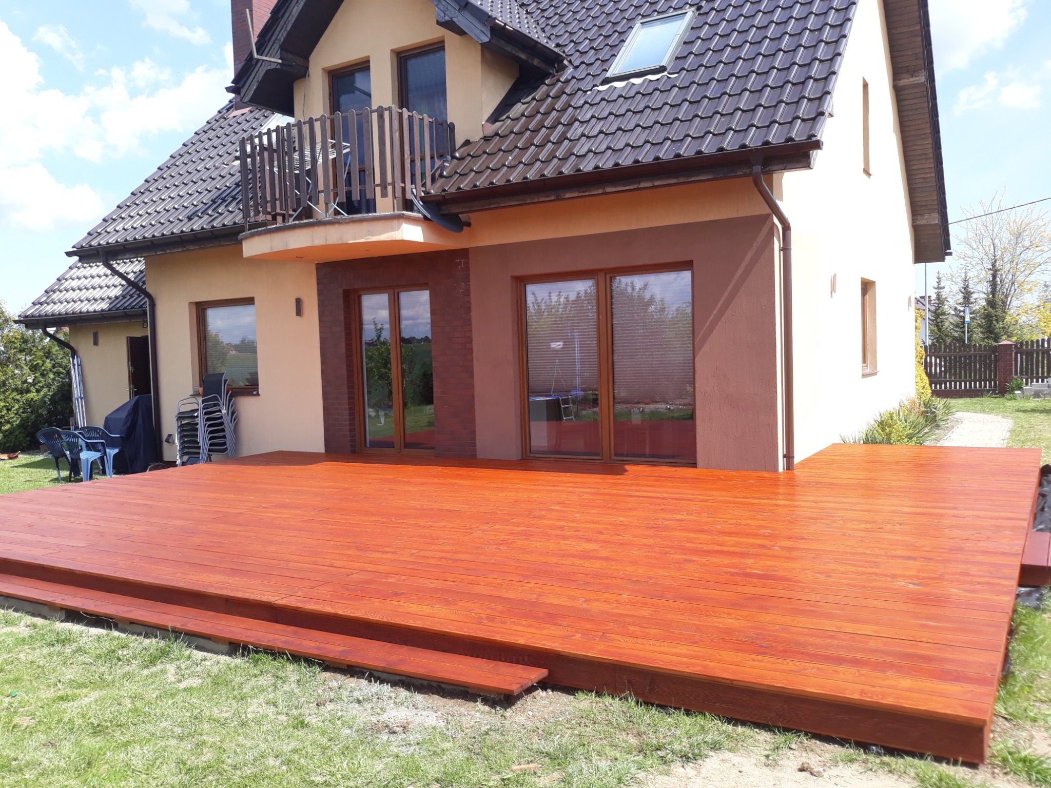 Nowoczesny taras drewniany z dwoma stopniami, wykonany z desek w kolorze ciepłego brązu, przylegający do domu z balkonem i dużymi oknami.