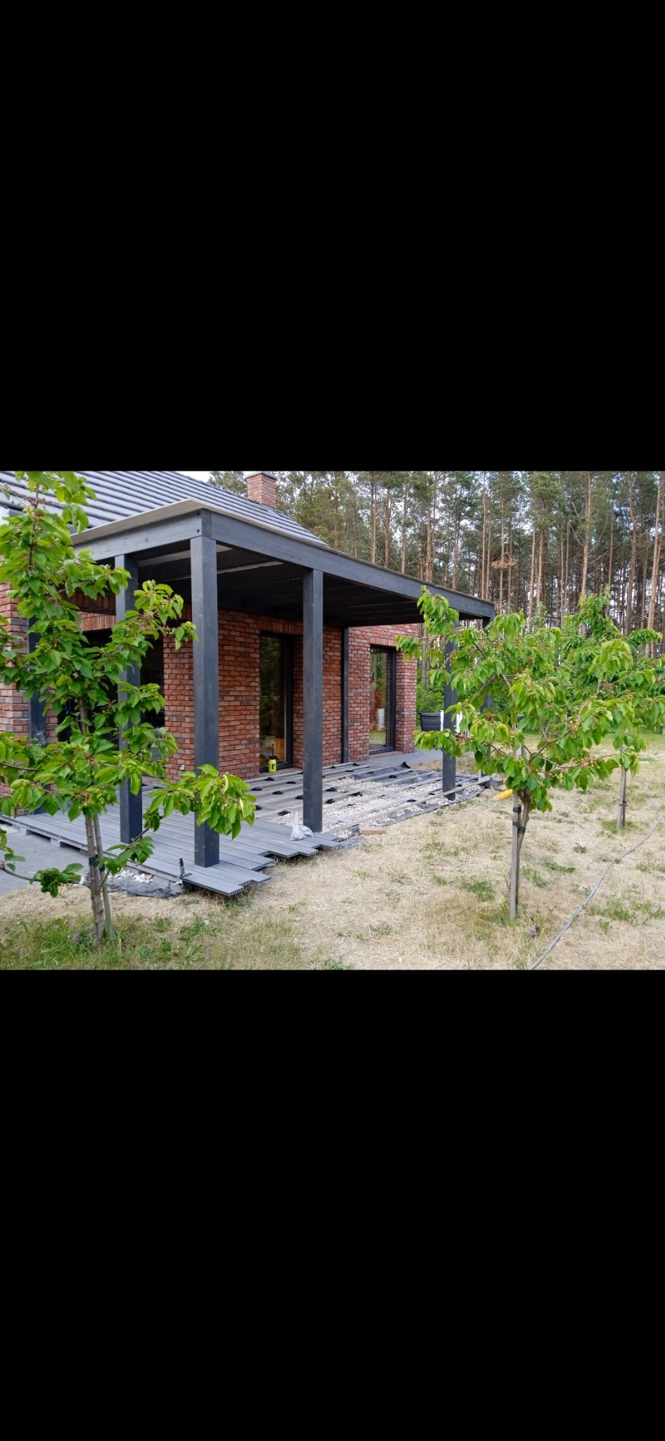 Montaż szarego tarasu z desek kompozytowych na podłożu z geowłókniny i żwiru, widoczne elementy konstrukcyjne pergoli z ciemnego drewna przy ceglanym budynku, w tle las.