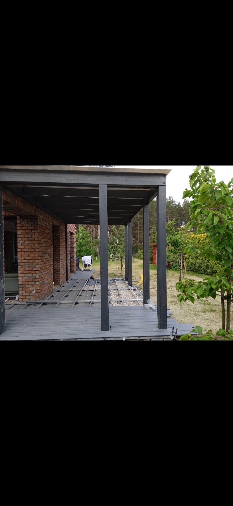 Budowa zadaszonego tarasu kompozytowego w trakcie realizacji: widoczne legary, częściowo ułożona deska, szara konstrukcja zadaszenia, ceglany budynek w tle.