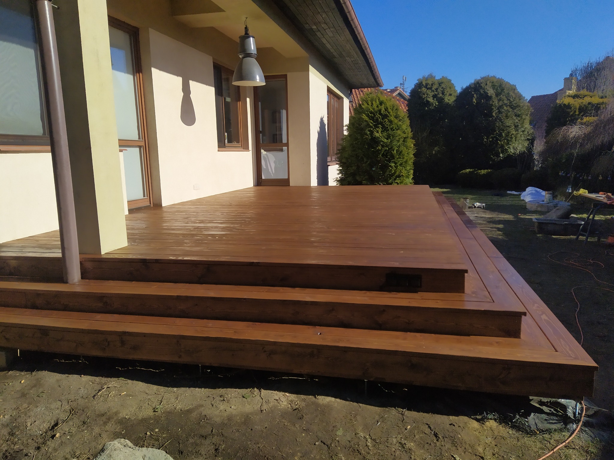 Brązowy taras z drewna z trzema stopniami prowadzącymi do domu, widok na ogród z zielonymi drzewami i krzewami, wisząca lampa na zewnątrz.