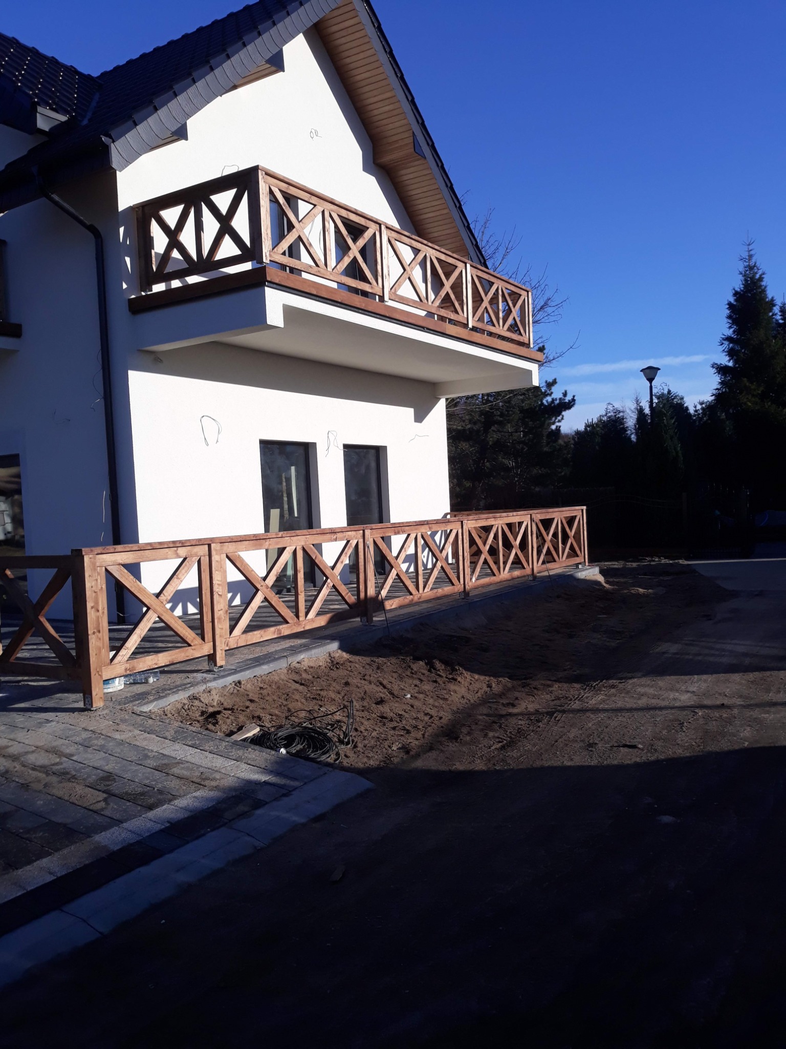 Nowoczesny dom z białymi ścianami i balkonem z drewnianą balustradą w kształcie krzyży, otoczony ziemią przygotowaną pod trawnik i niskim drewnianym ogrodzeniem o identycznym wzorze.