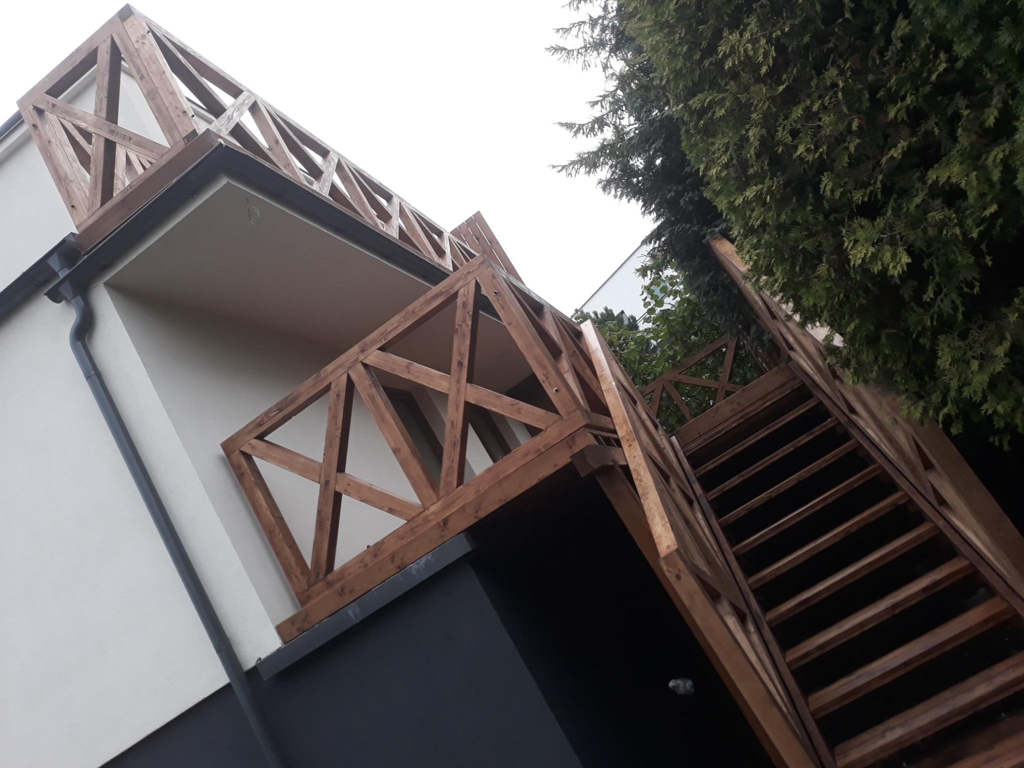 Kątowe ujęcie drewnianej konstrukcji schodów i balustrady tarasu z widocznym fragmentem budynku o białej elewacji i ciemnym cokole, całość na tle zieleni drzew.