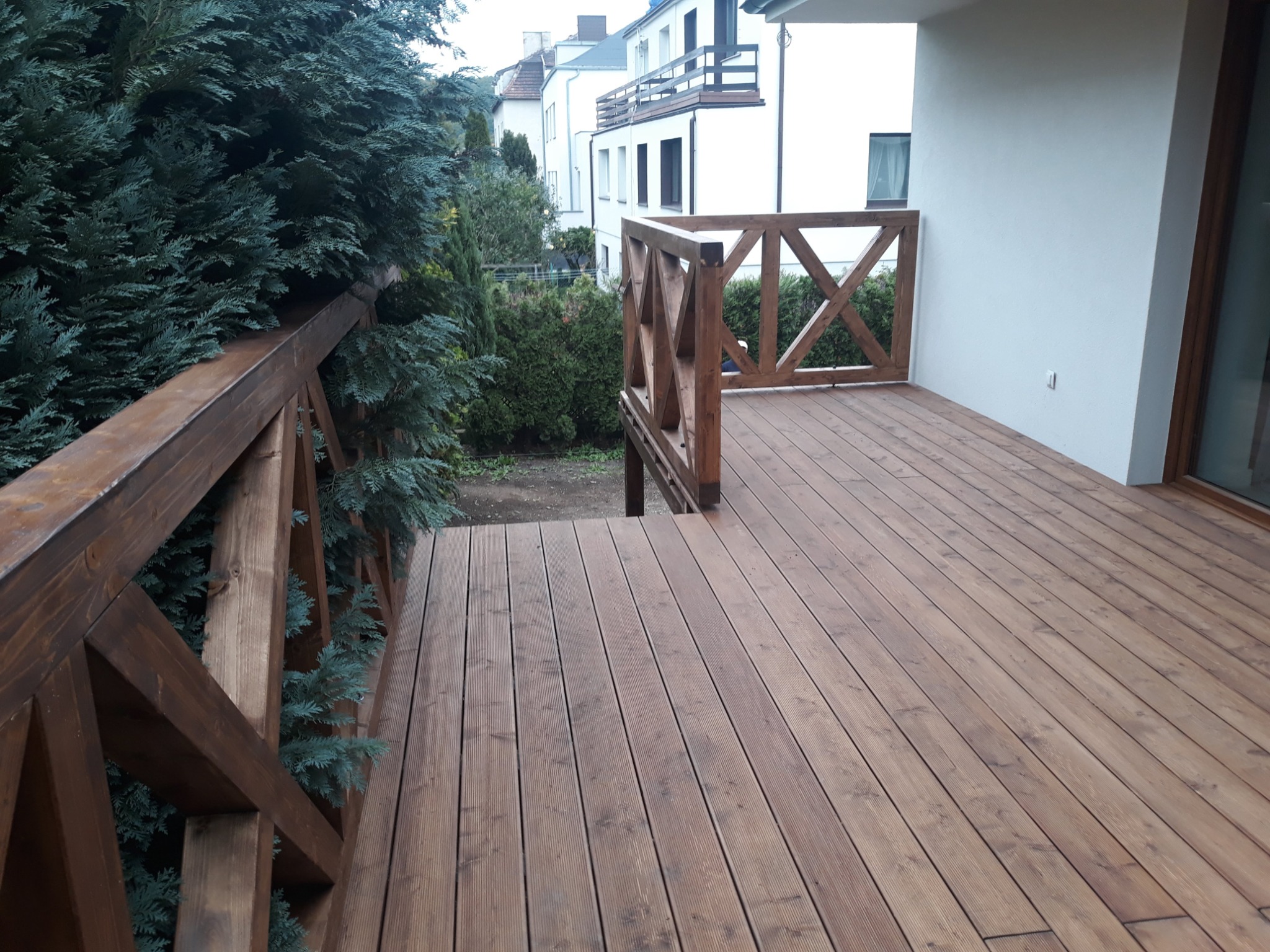 Drewniany taras z balustradą w stylu rustykalnym, widok z góry na deski i elementy konstrukcyjne, w tle zieleń i zabudowa mieszkalna.