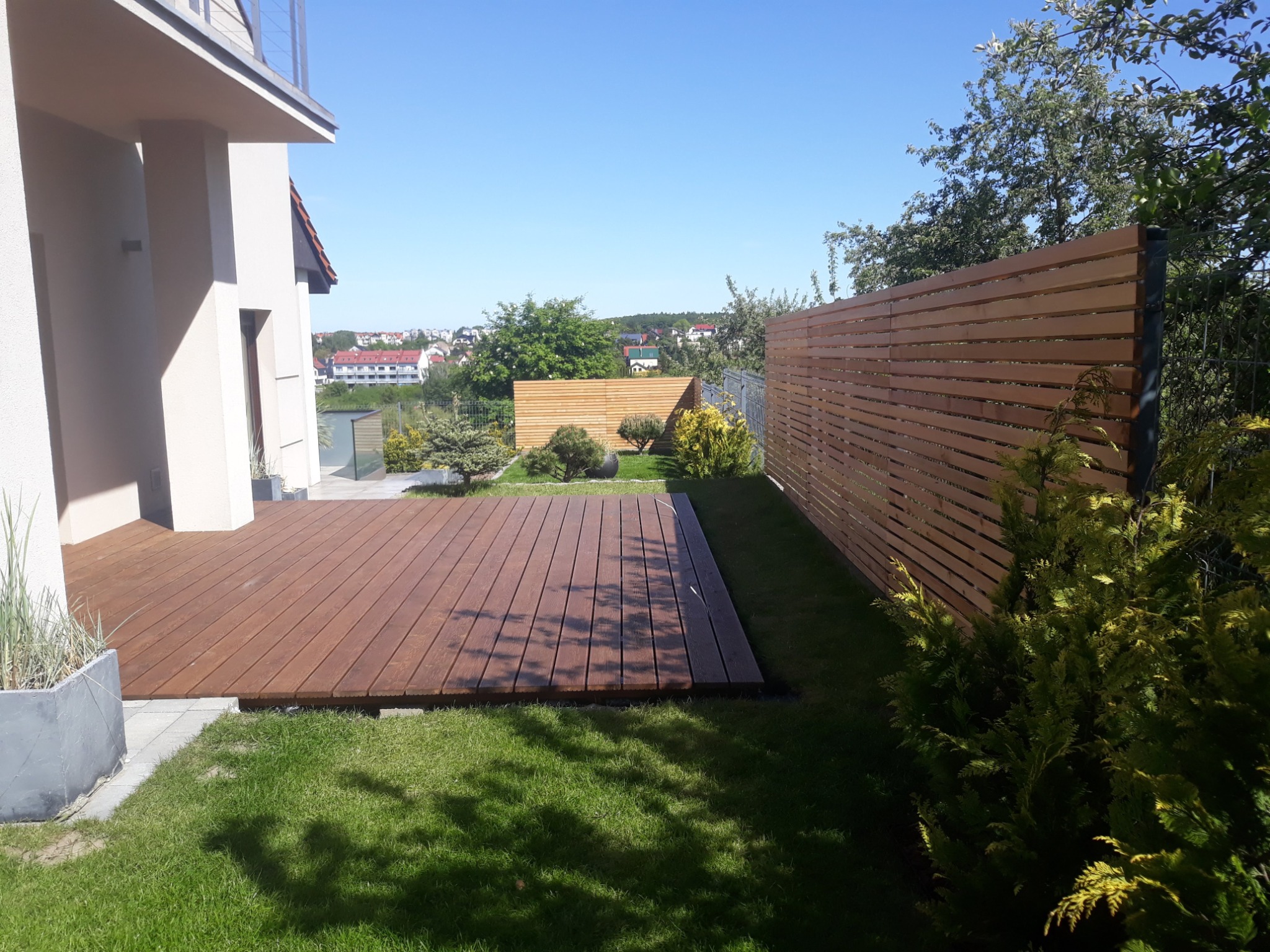 Nowoczesny taras z drewna egzotycznego przylegający do domu, otoczony trawnikiem i drewnianym ogrodzeniem lamelowym, w tle zabudowa miejska.