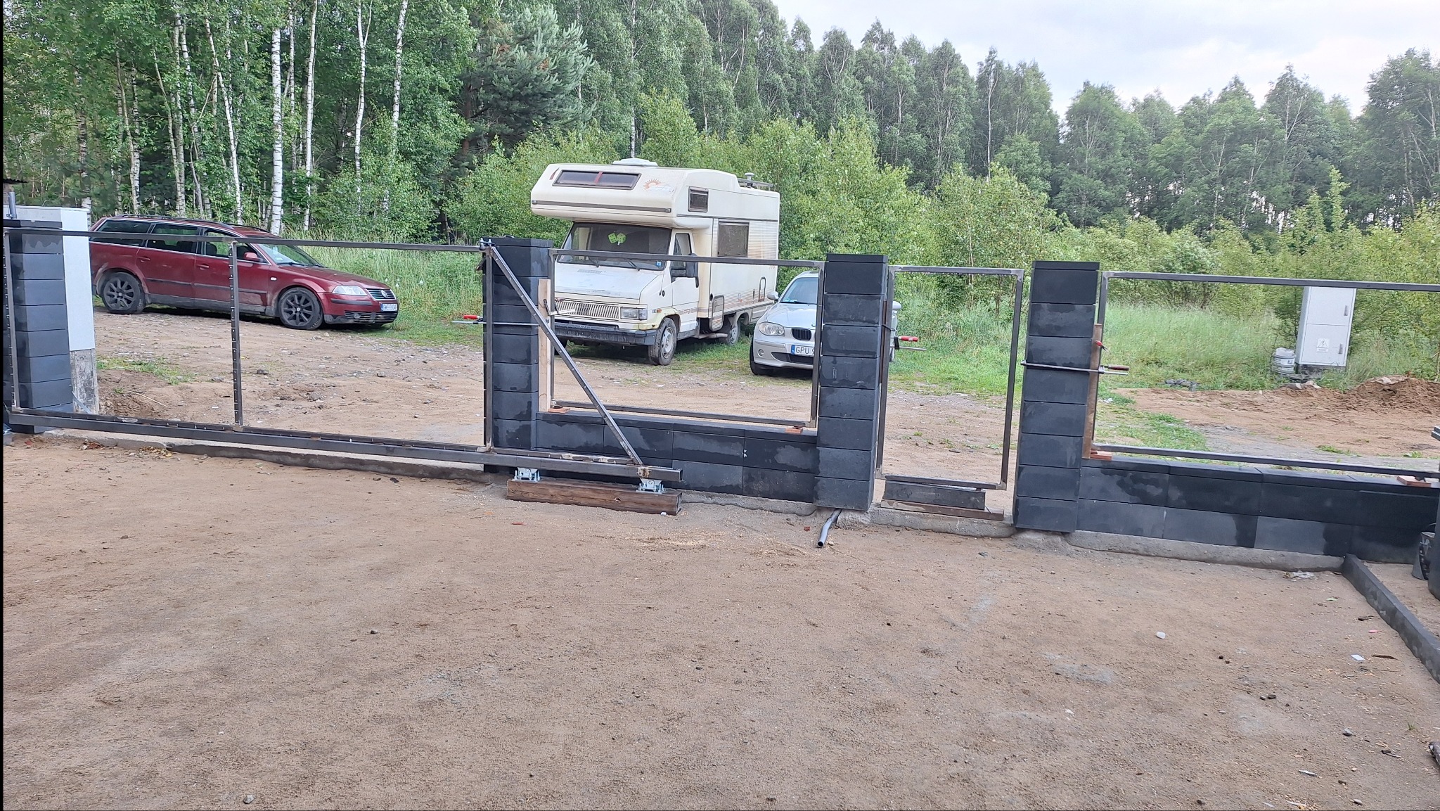 W trakcie budowy: brama wjazdowa z czarnych bloczków betonowych i metalowych elementów. W tle samochody osobowe i kamper na działce z drzewami.