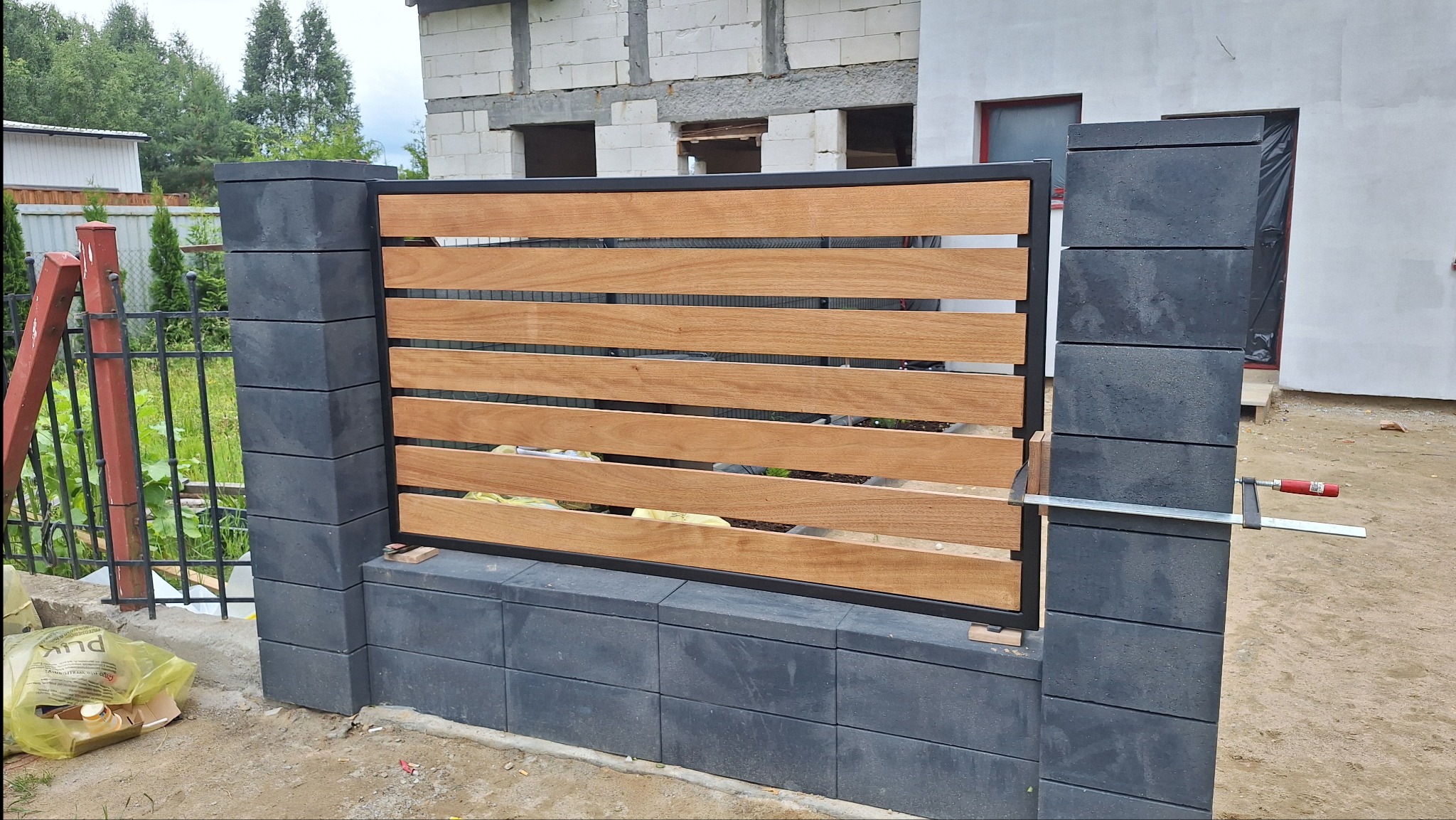 Nowoczesne przęsło ogrodzeniowe z poziomych desek i betonowych słupków w trakcie montażu, z widocznym ściskiem stolarskim. Surowy stan budowy w tle.