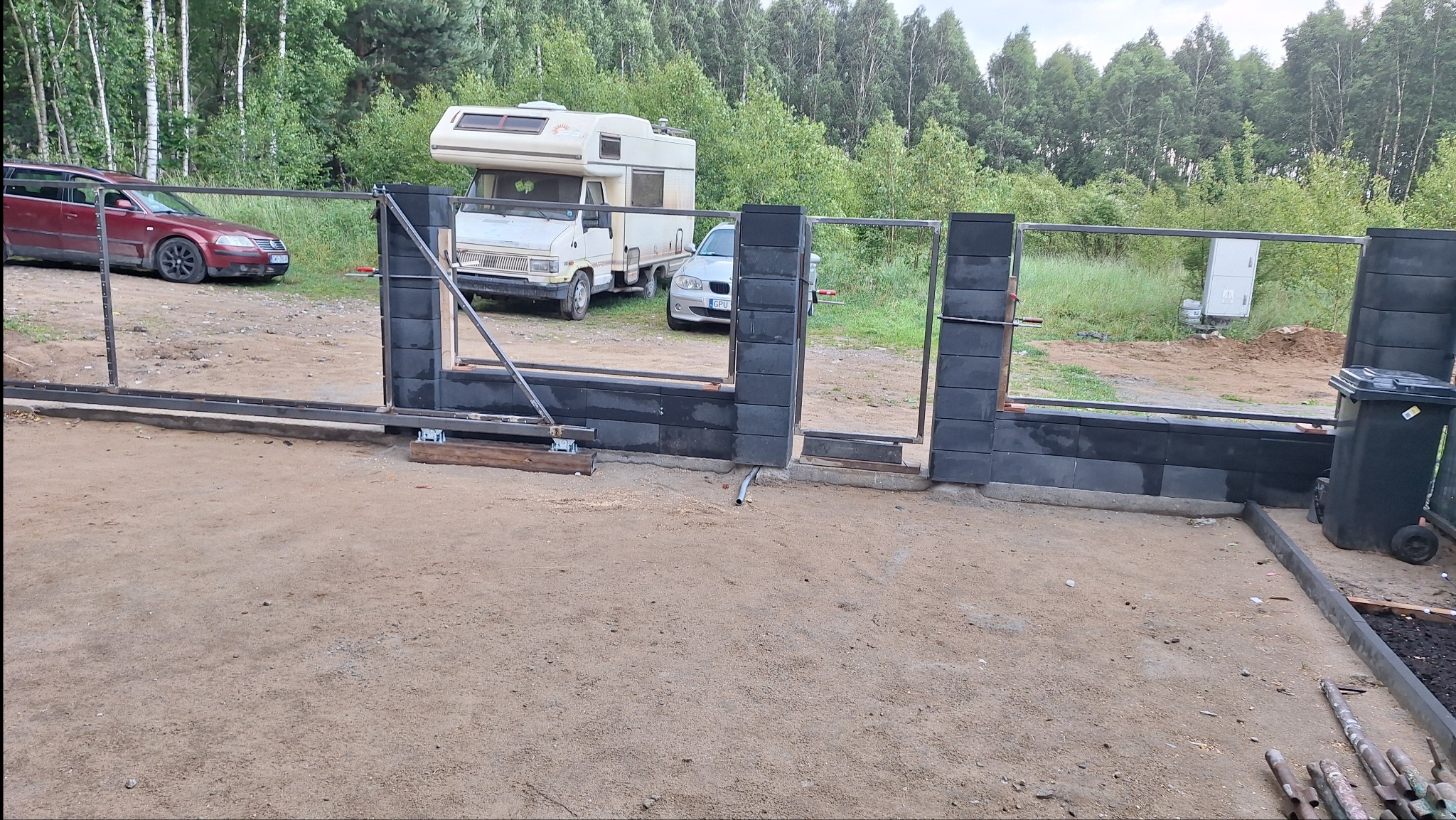 W trakcie budowy: fragment ogrodzenia z betonowych bloczków i metalowych elementów, widoczne samochody i zieleń w tle. Etap montażu, surowy wygląd.