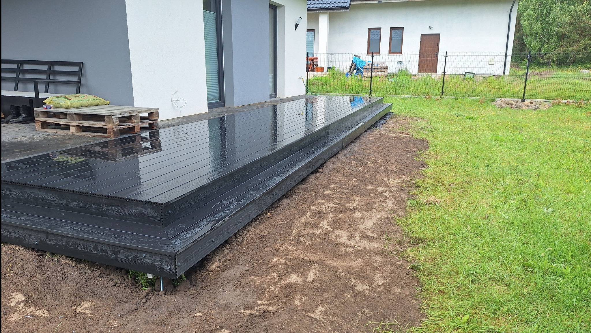 Nowoczesny taras kompozytowy w kolorze grafitowym, z widocznymi detalami montażu na metalowych wspornikach. Świeżo ułożony, z paletą i workiem w tle, na tle domu.