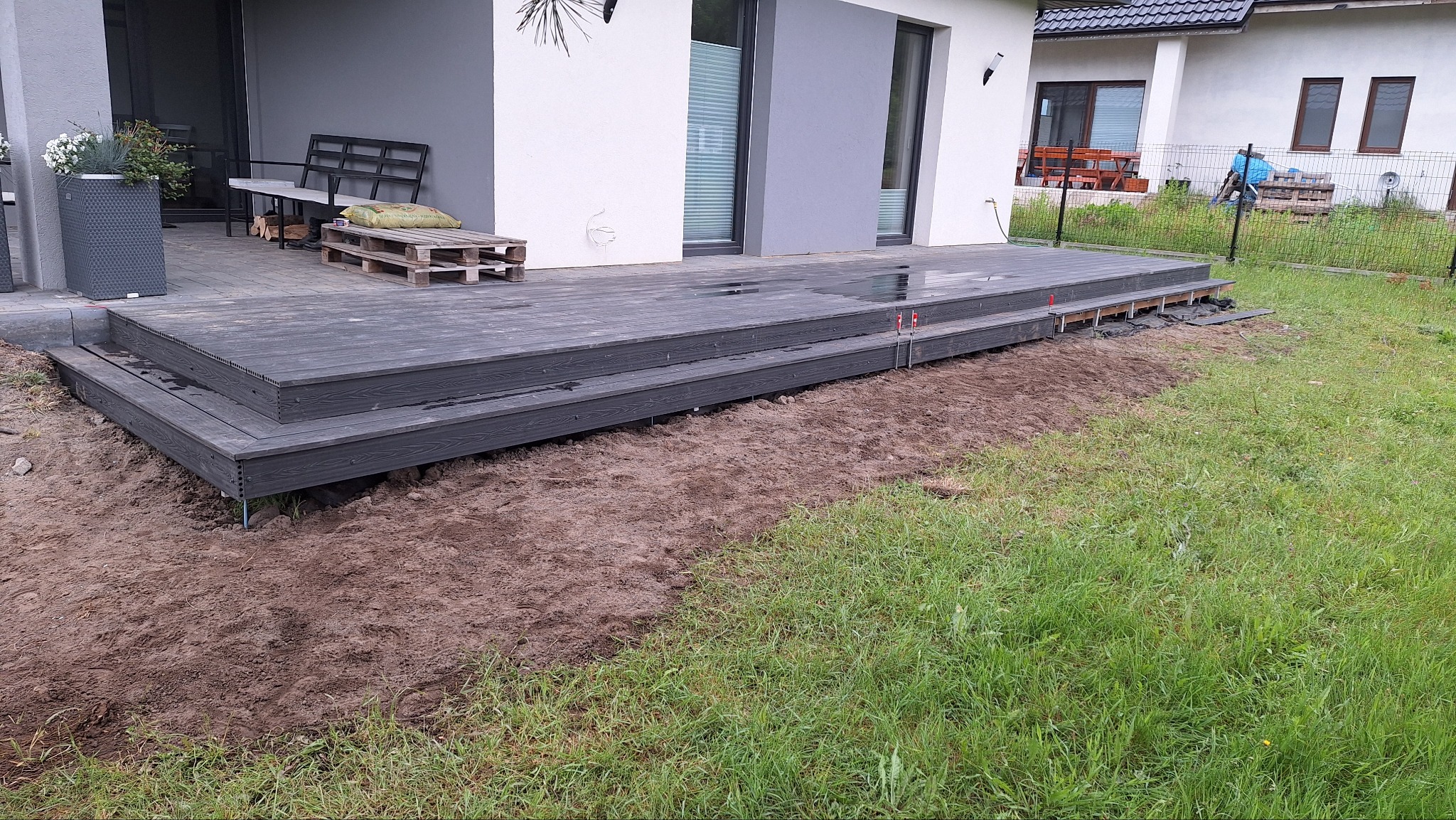 Nowoczesny taras kompozytowy z dwustopniowymi schodami, częściowo wykończony, z widocznym przygotowaniem terenu i elementami montażowymi. Minimalistyczny design.