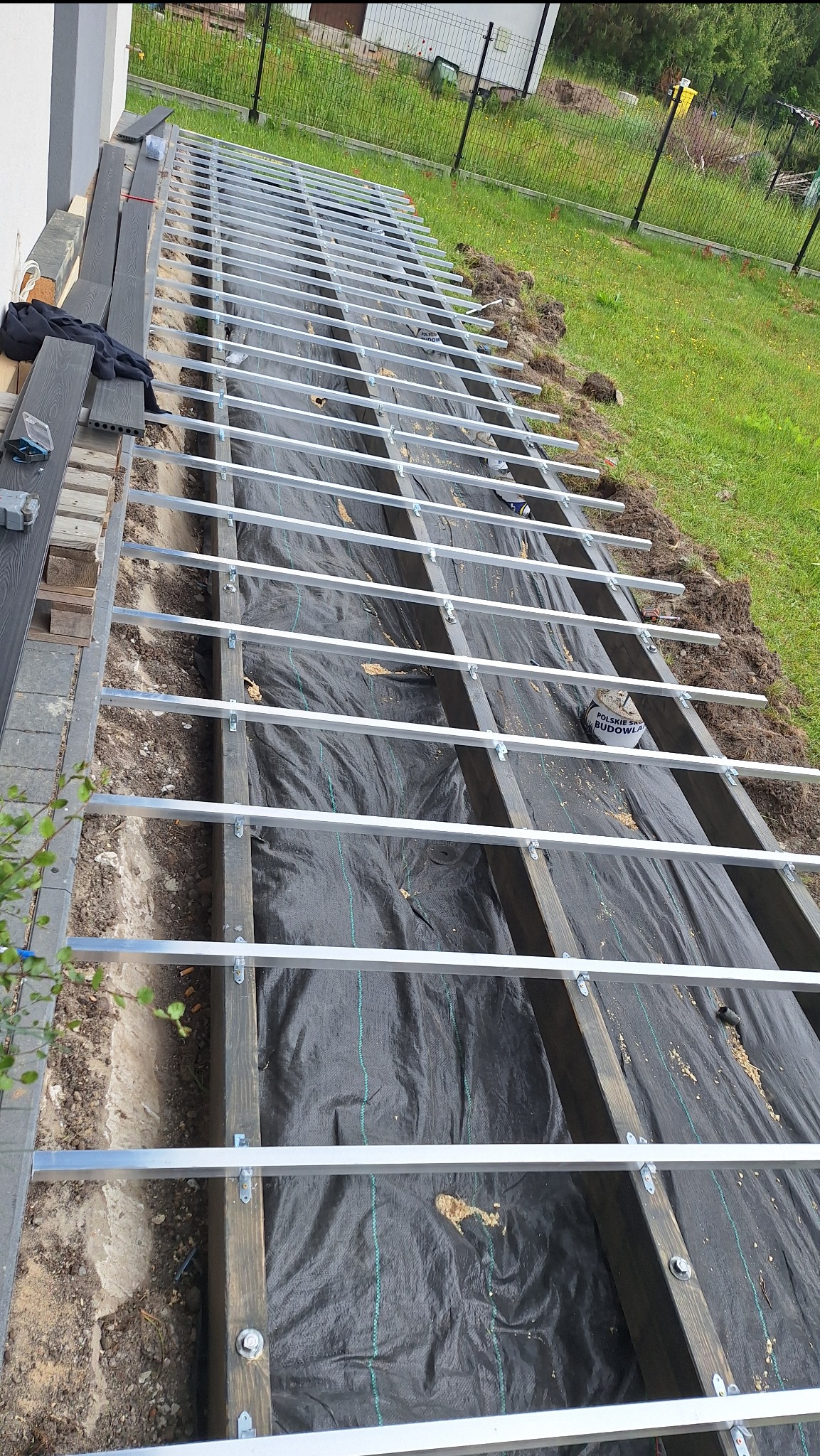 Konstrukcja tarasu w trakcie budowy: drewniane legary, metalowe poprzeczki, czarna geowłóknina. Częściowo ułożone deski tarasowe. Widok z góry, zieleń w tle.