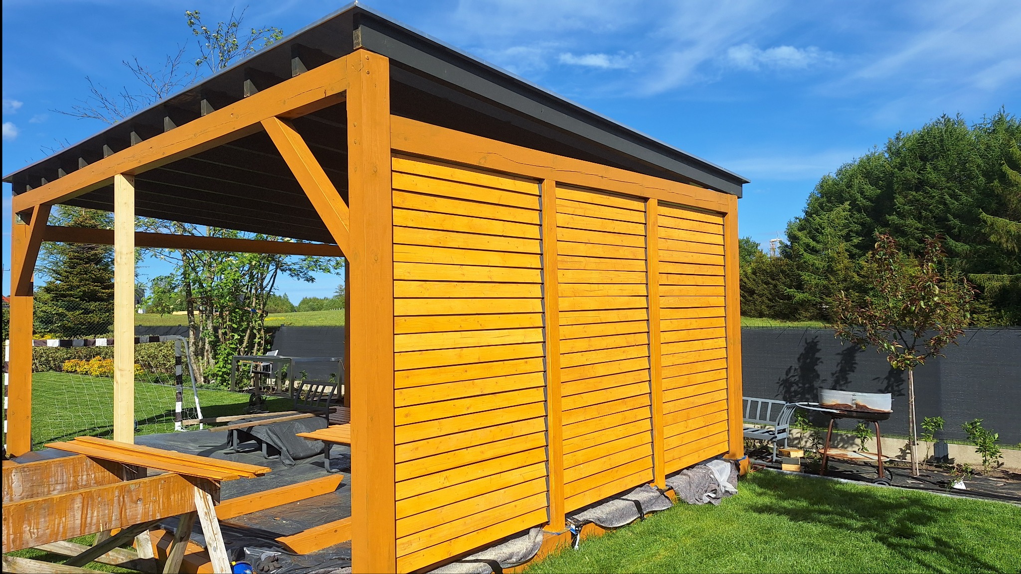 Drewniana altana z częściowo zabudowanymi ścianami, pomalowana na ciepły, miodowy kolor. Widoczne elementy konstrukcyjne i fragment ogrodu z grillem.