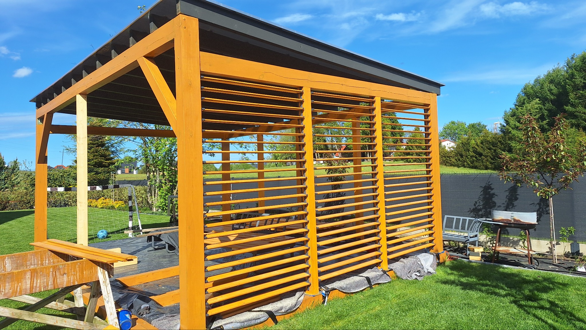Drewniana pergola z lamelami w ogrodzie, częściowo zmontowana. Widoczne narzędzia i materiały budowlane. W tle trawnik, drzewa i błękitne niebo.