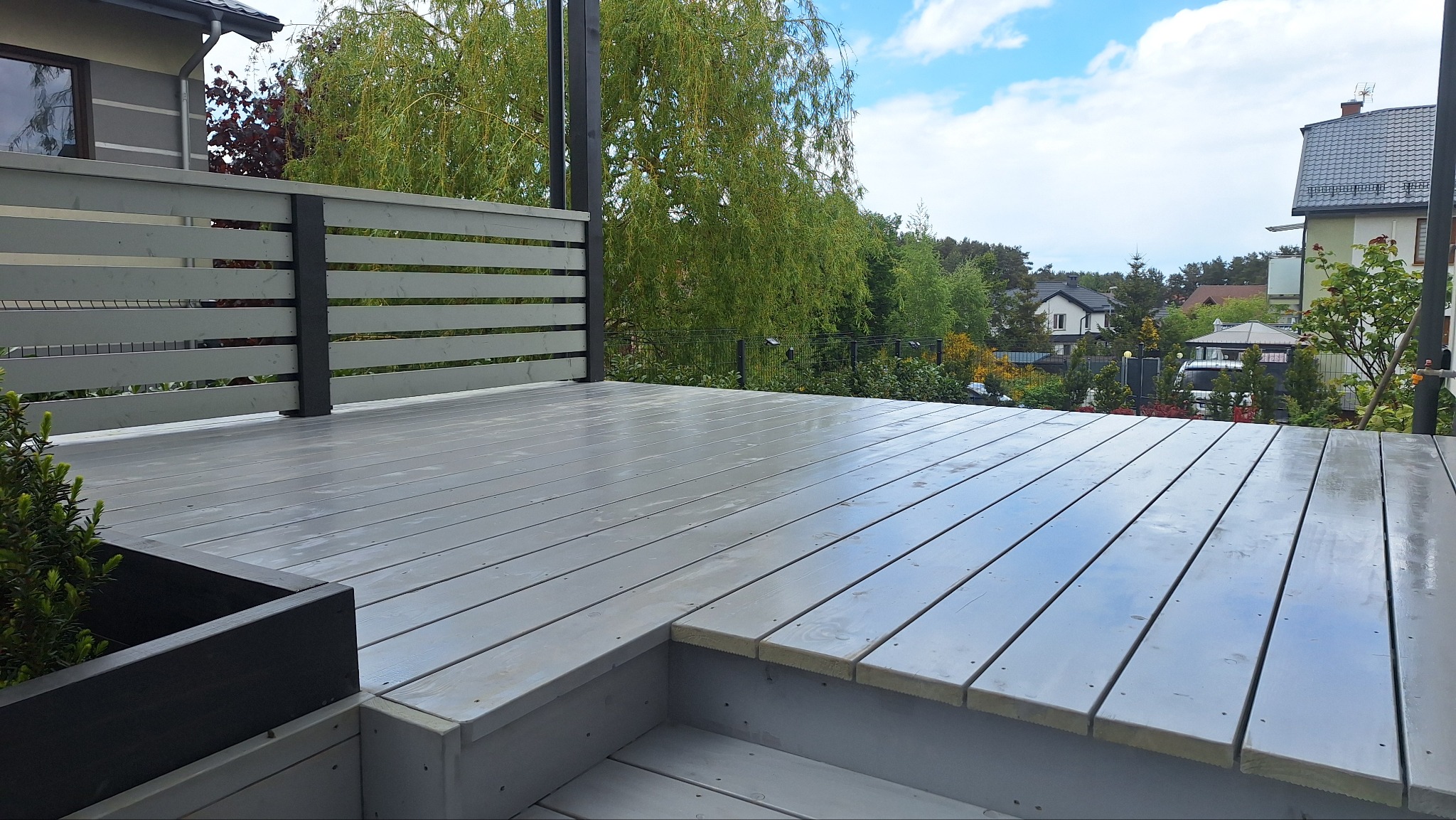 Szary taras drewniany z niskim, poziomym ogrodzeniem i schodami, otoczony zielenią. Widok na domy w tle pod błękitnym niebem z chmurami.