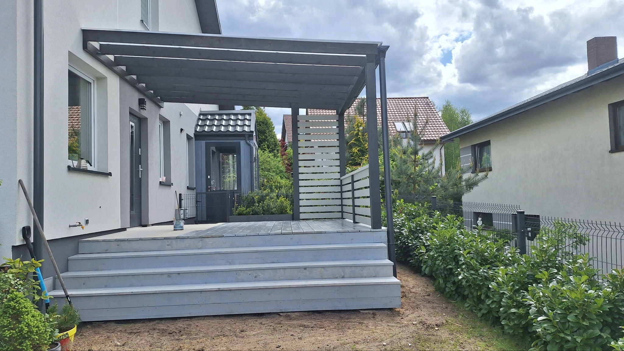 Szara pergola z drewnianym tarasem i schodami przy jasnym domu. Widoczne ogrodzenie panelowe i zieleń. Ujęcie z poziomu gruntu, pochmurne niebo w tle.