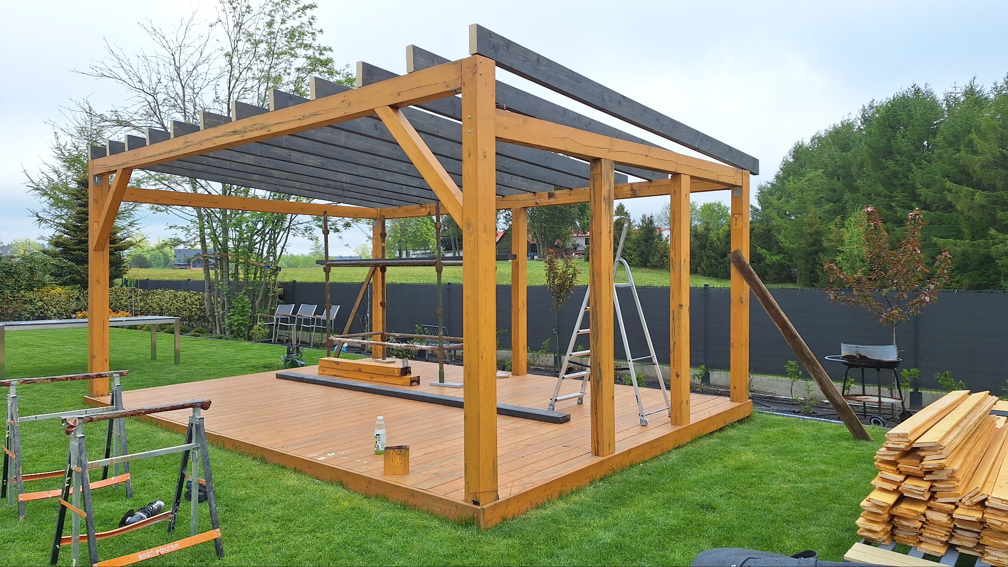 Drewniana pergola w trakcie budowy na tarasie. Widoczne elementy konstrukcyjne, drabina i narzędzia. Trawa, drzewa i czarne ogrodzenie w tle.